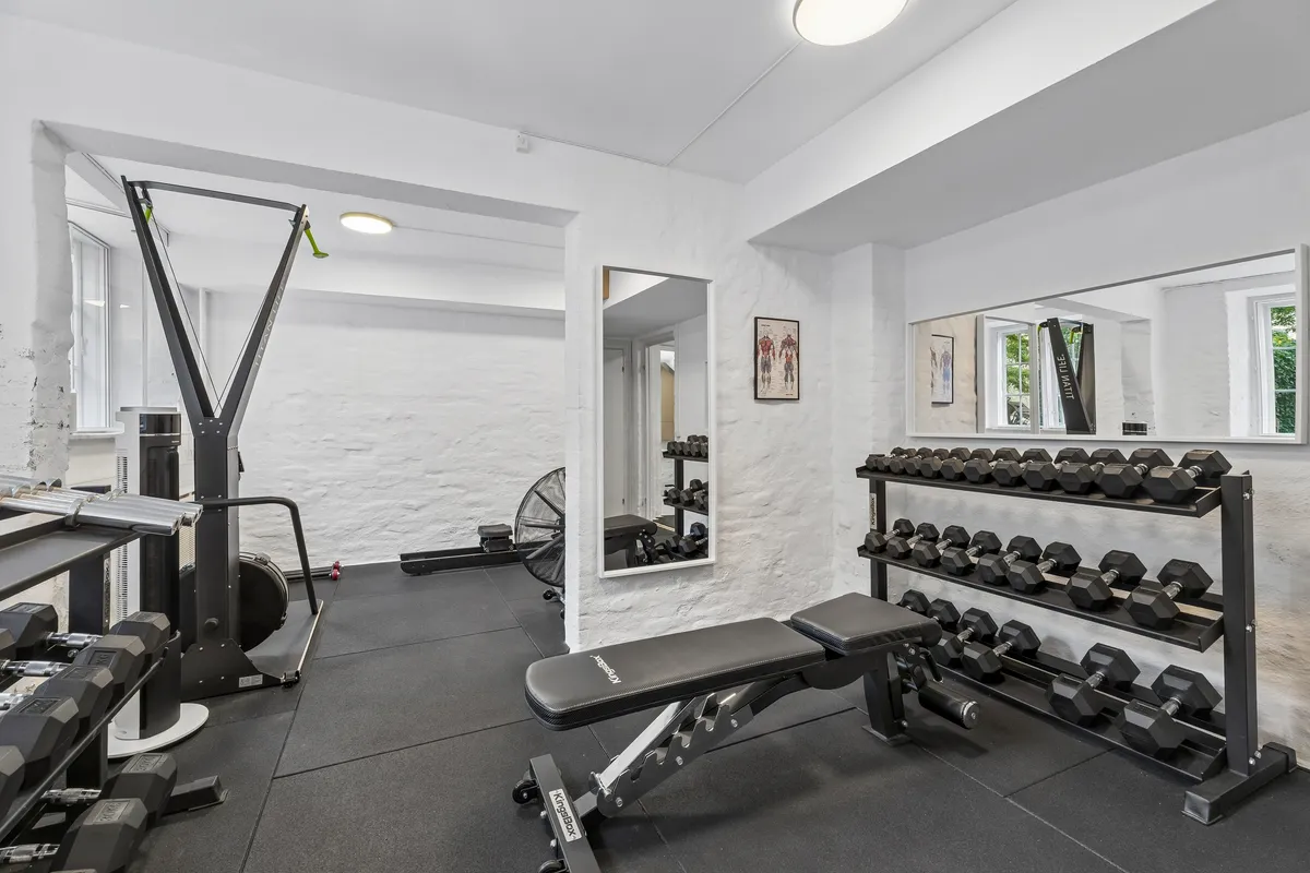 Fitnessrum med træningsapparater og vægte. Fladt loft og hvide vægge. Vinduer med udsigt til gårdsplads. Træningsbænk og håndvægte i sort og gråt
