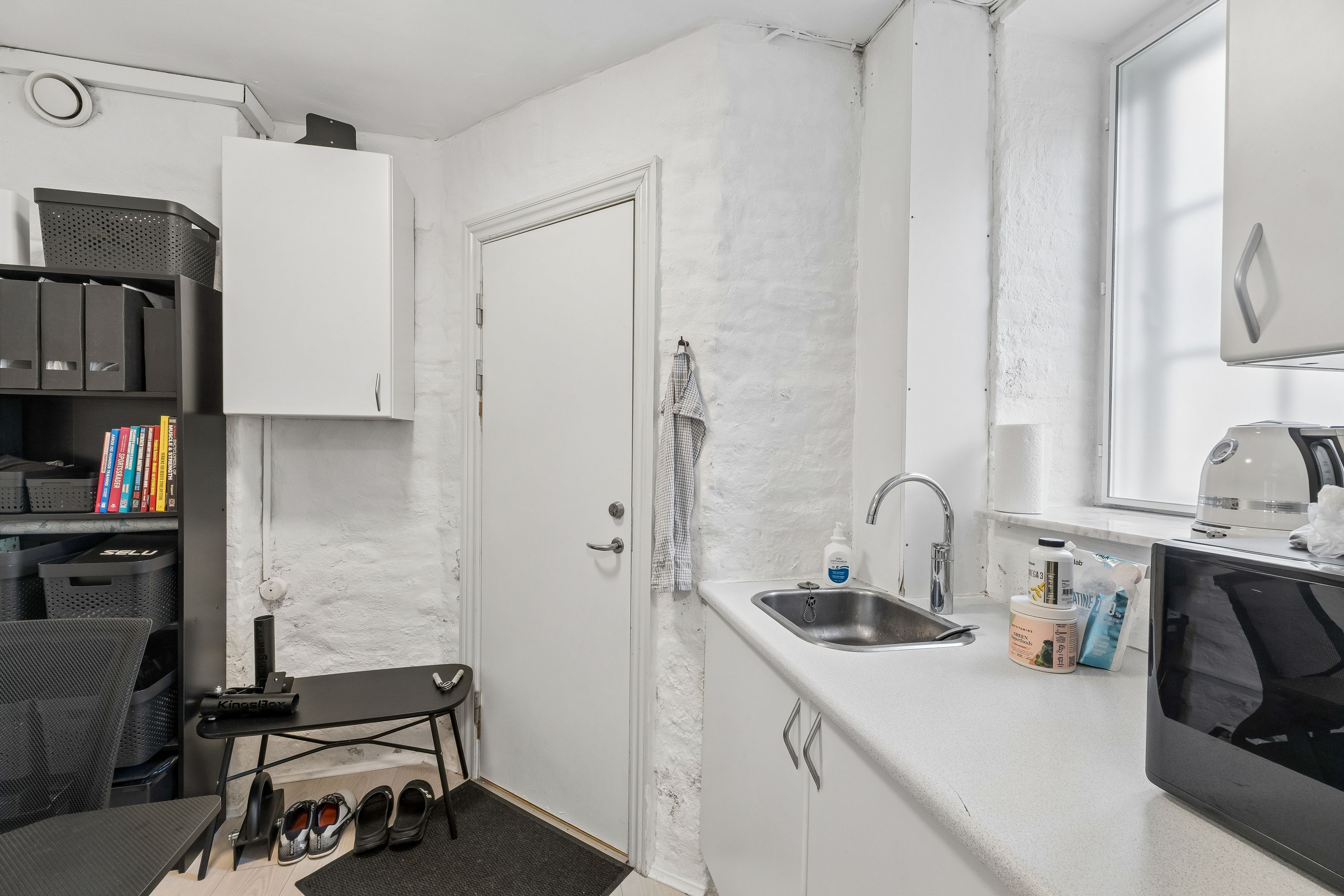 Lejlighed med lyse gulve og hvidt køkken med hætte over ovnen. Væggen er hvid og gul, og der er en stor, hvid skab. En stol og en stol står på gulvet.