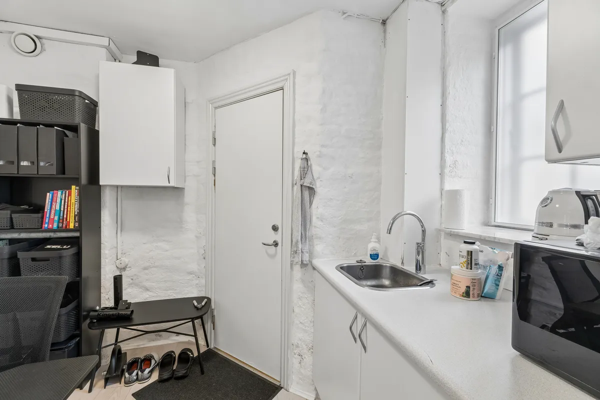 Lejlighed med lyse gulve og hvidt køkken med hætte over ovnen. Væggen er hvid og gul, og der er en stor, hvid skab. En stol og en stol står på gulvet.