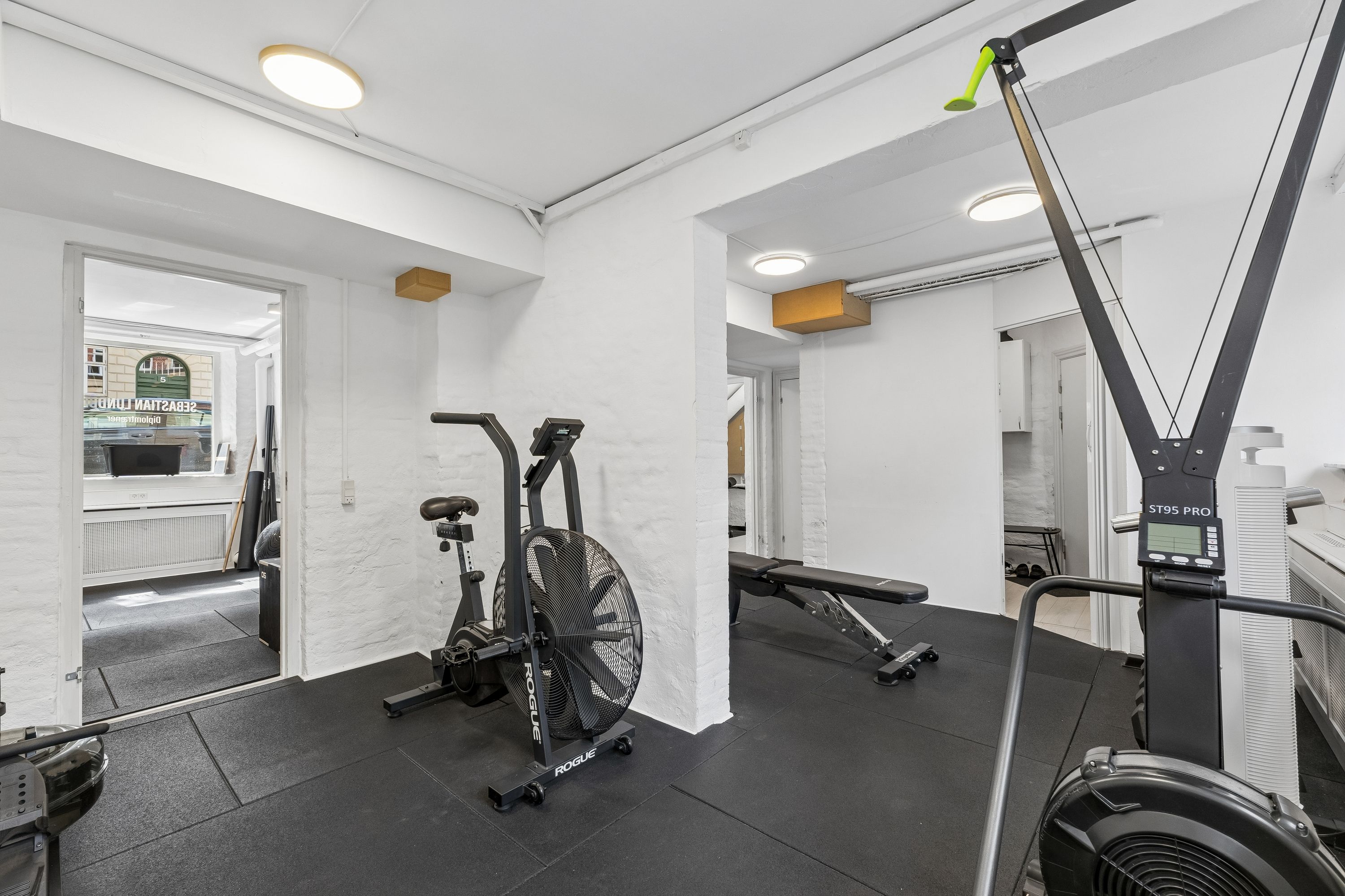 En fitnesslokale med håndcykel, løbemaskine, træningsbænk og træningstryk. Lyse lofter og moderne træningsudstyr.