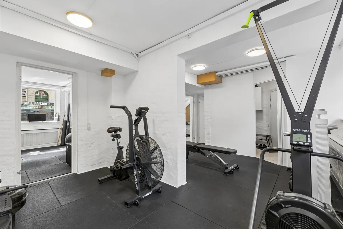En fitnesslokale med håndcykel, løbemaskine, træningsbænk og træningstryk. Lyse lofter og moderne træningsudstyr.