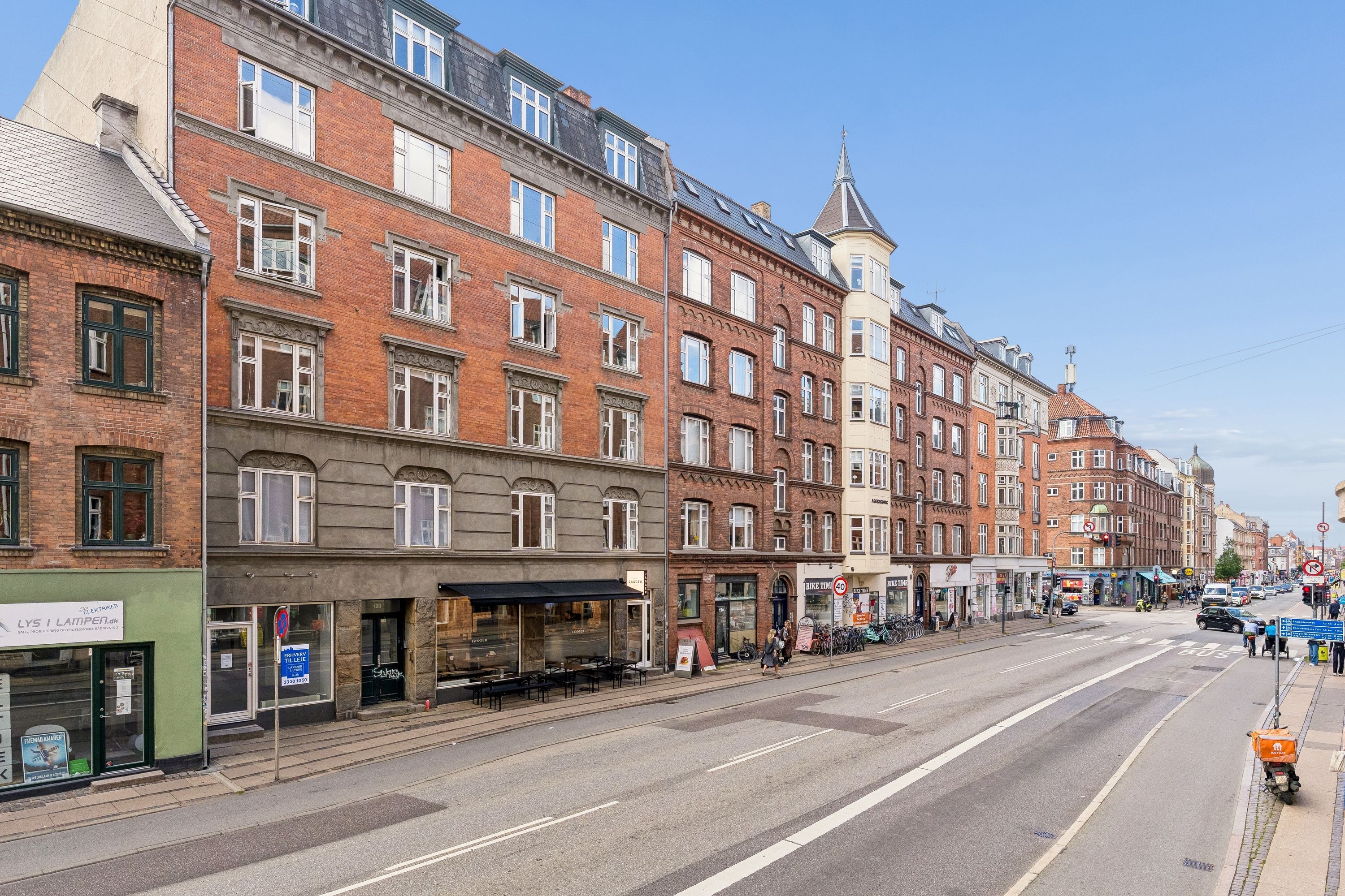 Store bygninger med mange vinduer og terrasser. To personer går på fortovet med en barnevogn. Biler og folk kører på en stor gade. En cykelstativ med mange cykler. En stor bygning med en spids tag.
