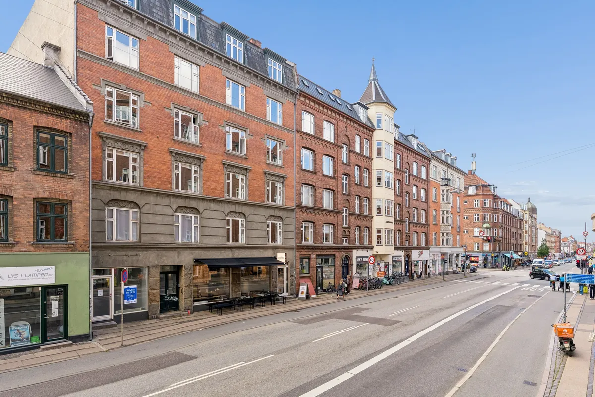 Store bygninger med mange vinduer og terrasser. To personer går på fortovet med en barnevogn. Biler og folk kører på en stor gade. En cykelstativ med mange cykler. En stor bygning med en spids tag.