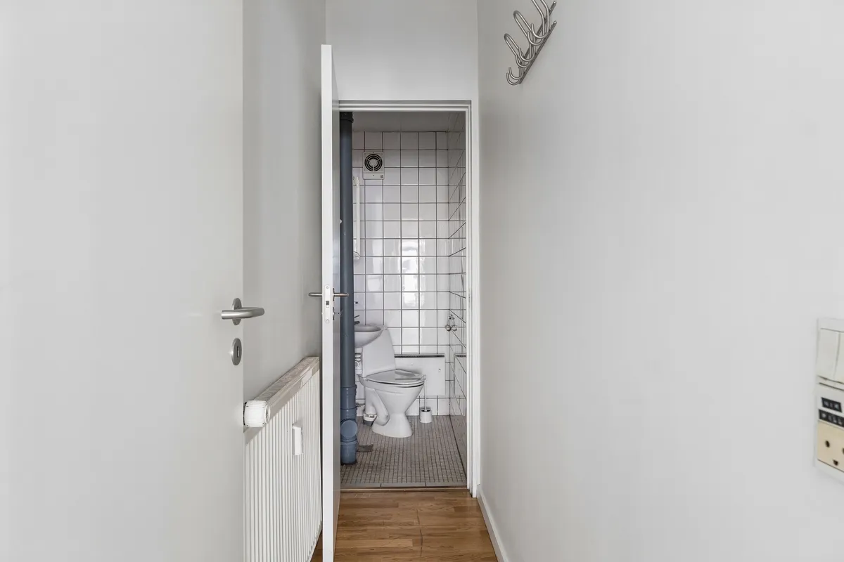 Toilet med hvidt tøjskab, toilet og vandhane i en lille, lyse stue med hvidt klinkerede vægge og trægulv