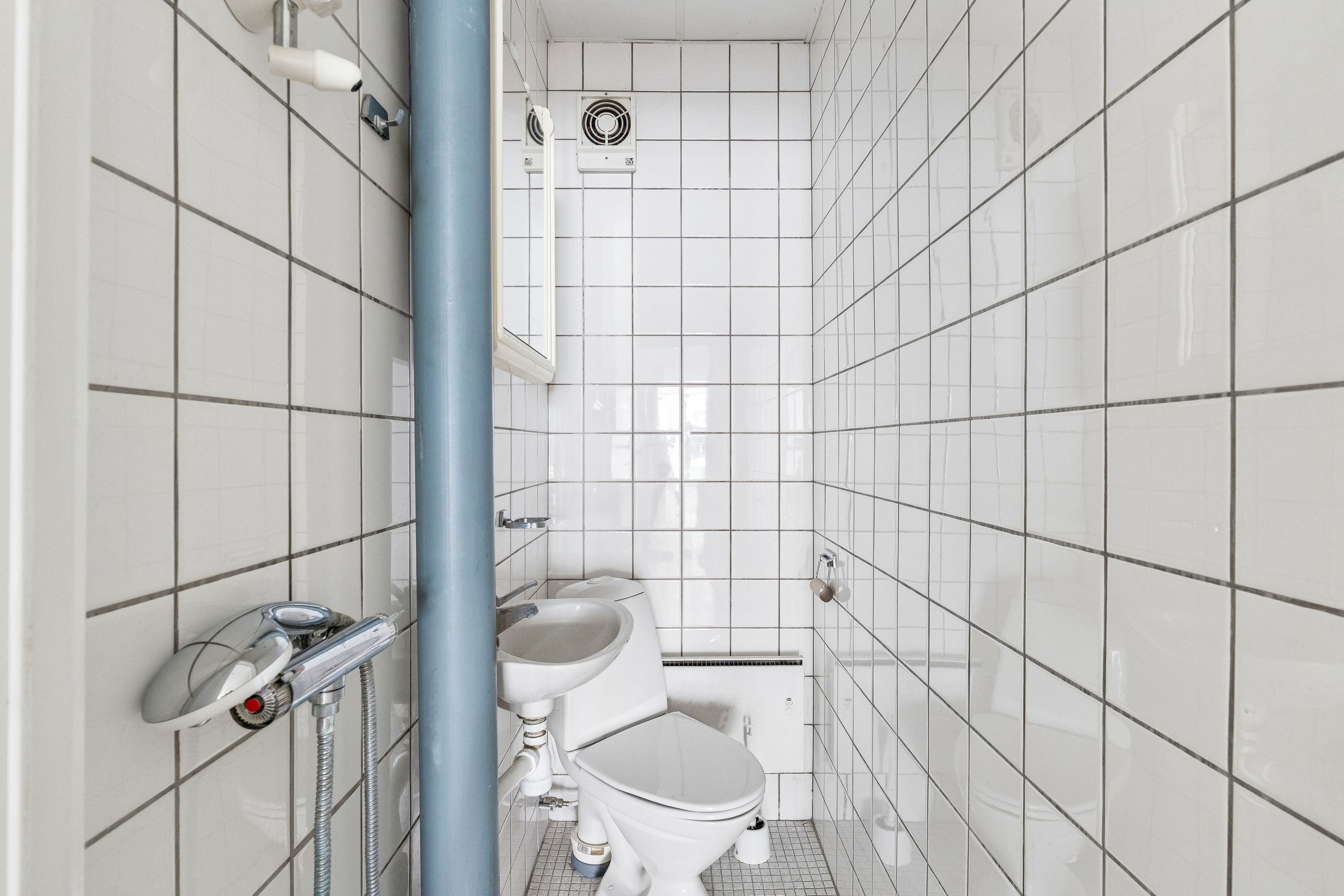En lille, hvidt flisdeklædt toiletrum med hvidt toilet og hvidt vask. Der er en håndholdt brusehoved og en ventileret loftsventil i rummet