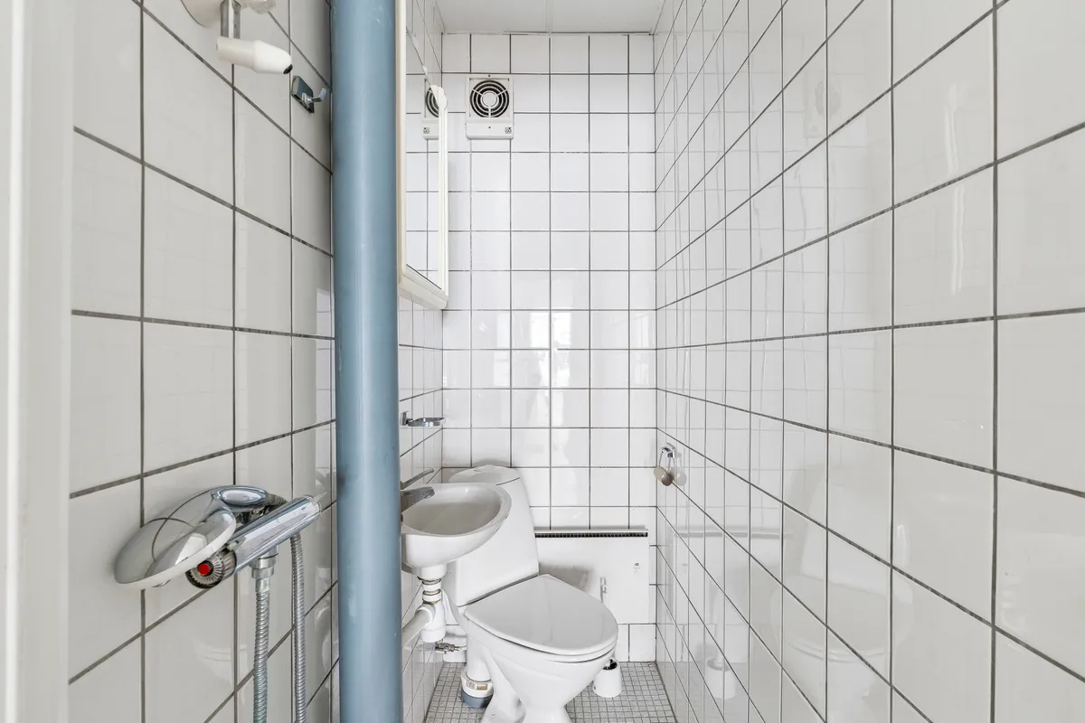 En lille, hvidt flisdeklædt toiletrum med hvidt toilet og hvidt vask. Der er en håndholdt brusehoved og en ventileret loftsventil i rummet
