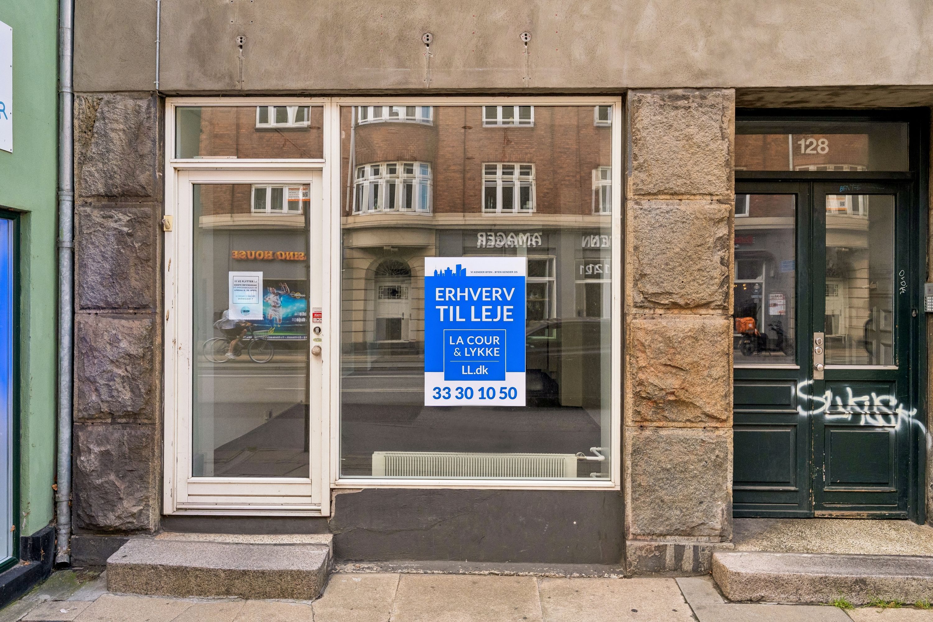 Butikstilbud på Amagerbrogade 128 i København S med skilt og glasdør, udvendig refleksion af cyklist og bil, indvendig varmeapparat og grafisk annonce