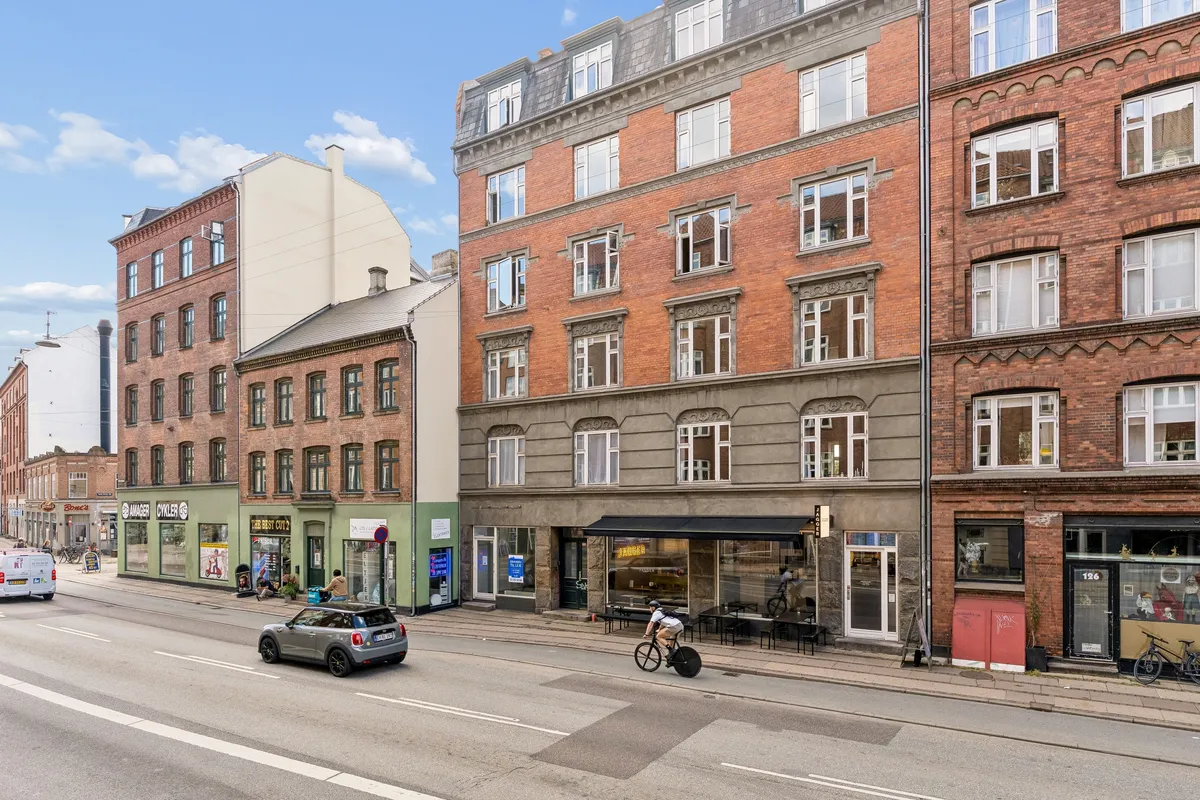 Butik til leje på Amagerbrogade med røde mursten og store ruder i 2300 København S. En person cykler på fortovet foran butikken, mens en bil passerer på gaden