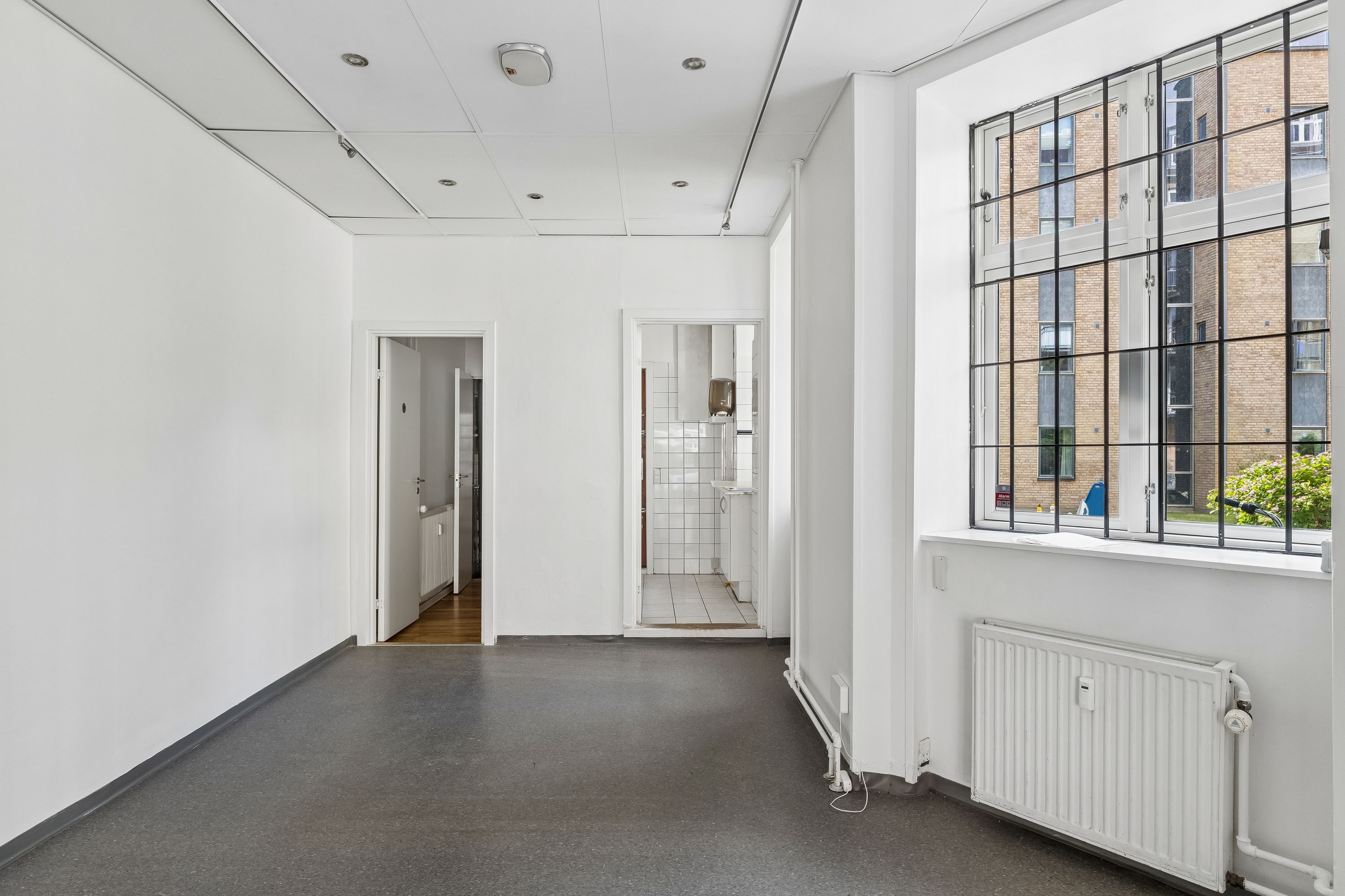 Minimalistisk og rummelig butikslokal til leje med hvide vægge, sort gulv, radiator og store vinduer til Amagerbrogade. Der er to adgangsdøre, og i baggrunden ses et bygningsanlæg med røde mursten.