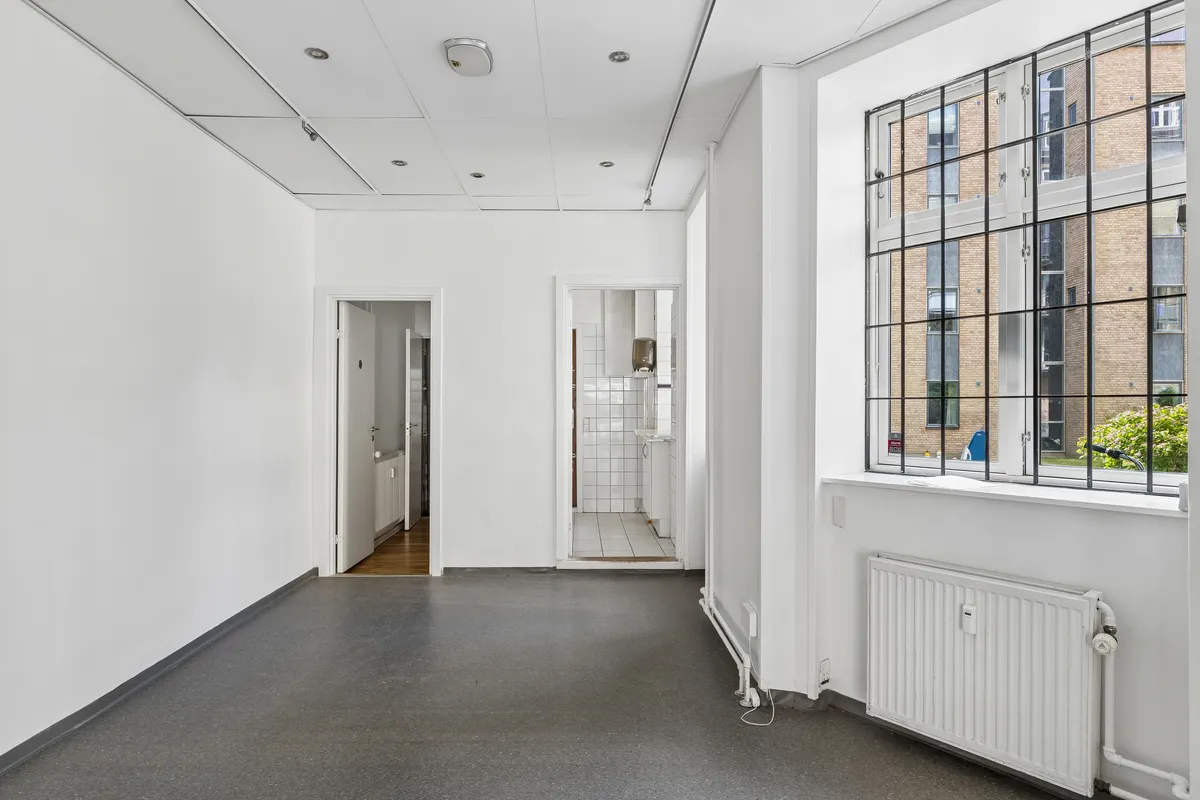 Minimalistisk og rummelig butikslokal til leje med hvide vægge, sort gulv, radiator og store vinduer til Amagerbrogade. Der er to adgangsdøre, og i baggrunden ses et bygningsanlæg med røde mursten.