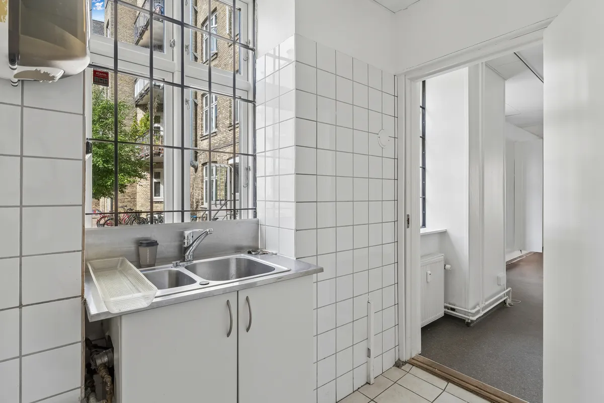 Rent og nyt køkken med lyse, hvide fliser, to nye rustfrit stål-vaskebædre, moderne lysekroner og udsigt til en søjle.
