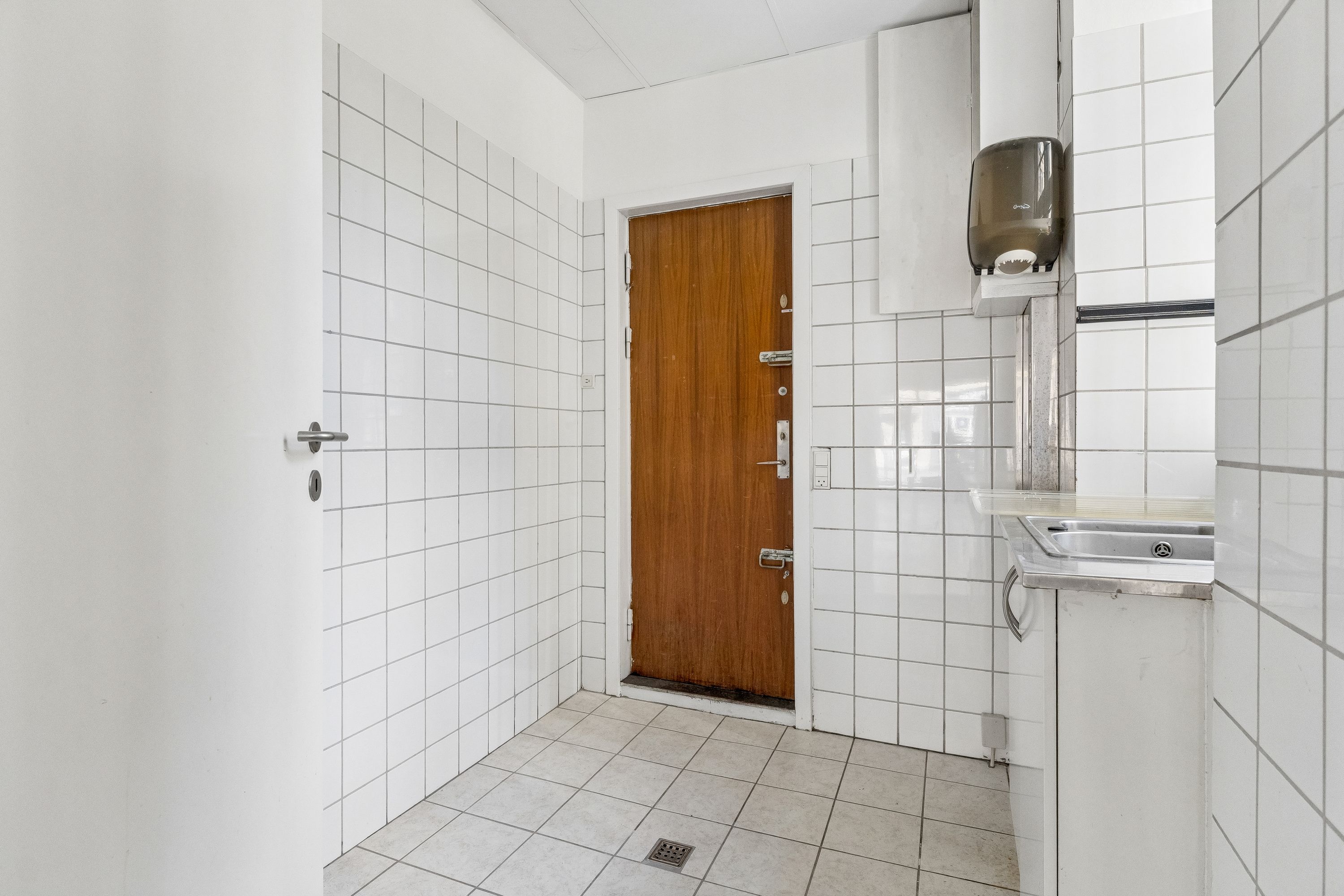 Rent og pænt badeværelse med hvidt fliser og møbler, to toiletpapirholdere og en håndvask med spejl. Væggene er hvide og der er et lukket trædør og en dørknop
