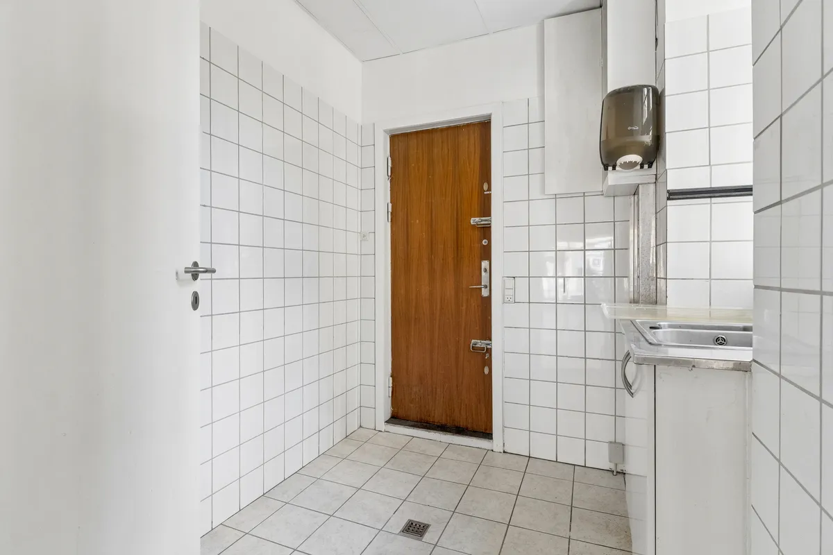 Rent og pænt badeværelse med hvidt fliser og møbler, to toiletpapirholdere og en håndvask med spejl. Væggene er hvide og der er et lukket trædør og en dørknop