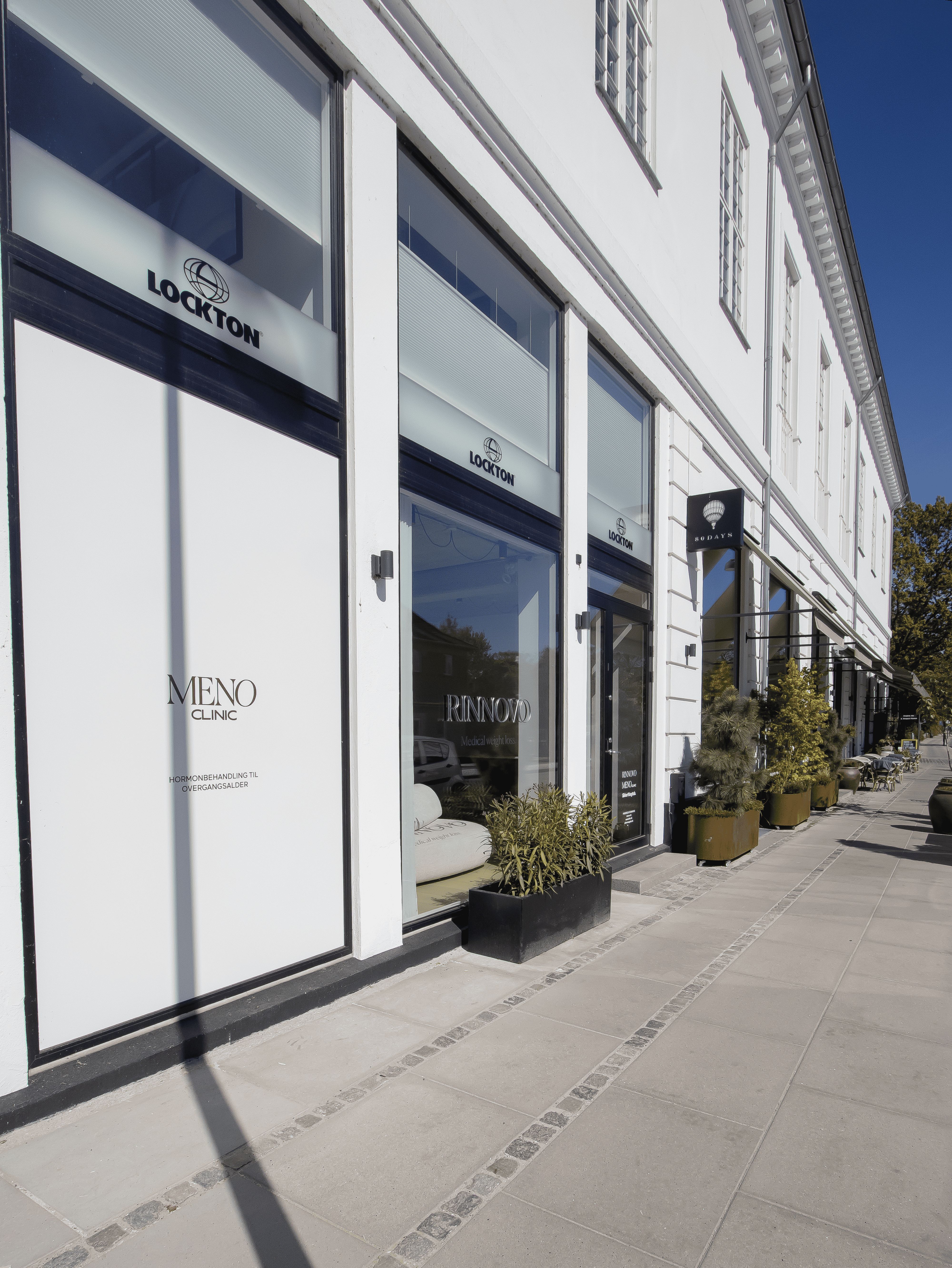 Meno Clinic ligger i et hvidt bygningshotel i Hellerup, med glasvinduer og røde vinduer. Planter og møbler er placeret udenfor, og der er flere butikker langs gaden