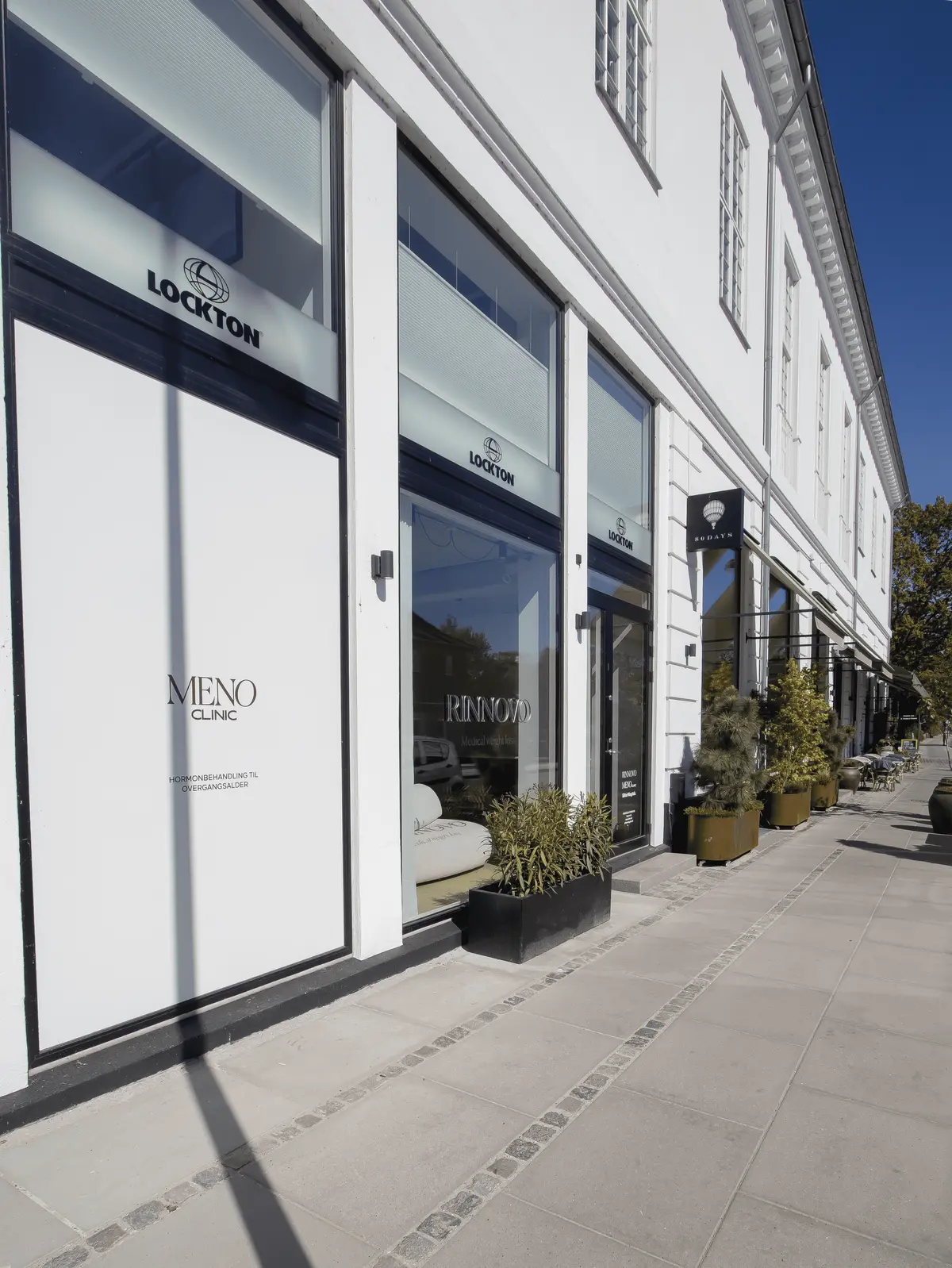Meno Clinic ligger i et hvidt bygningshotel i Hellerup, med glasvinduer og røde vinduer. Planter og møbler er placeret udenfor, og der er flere butikker langs gaden