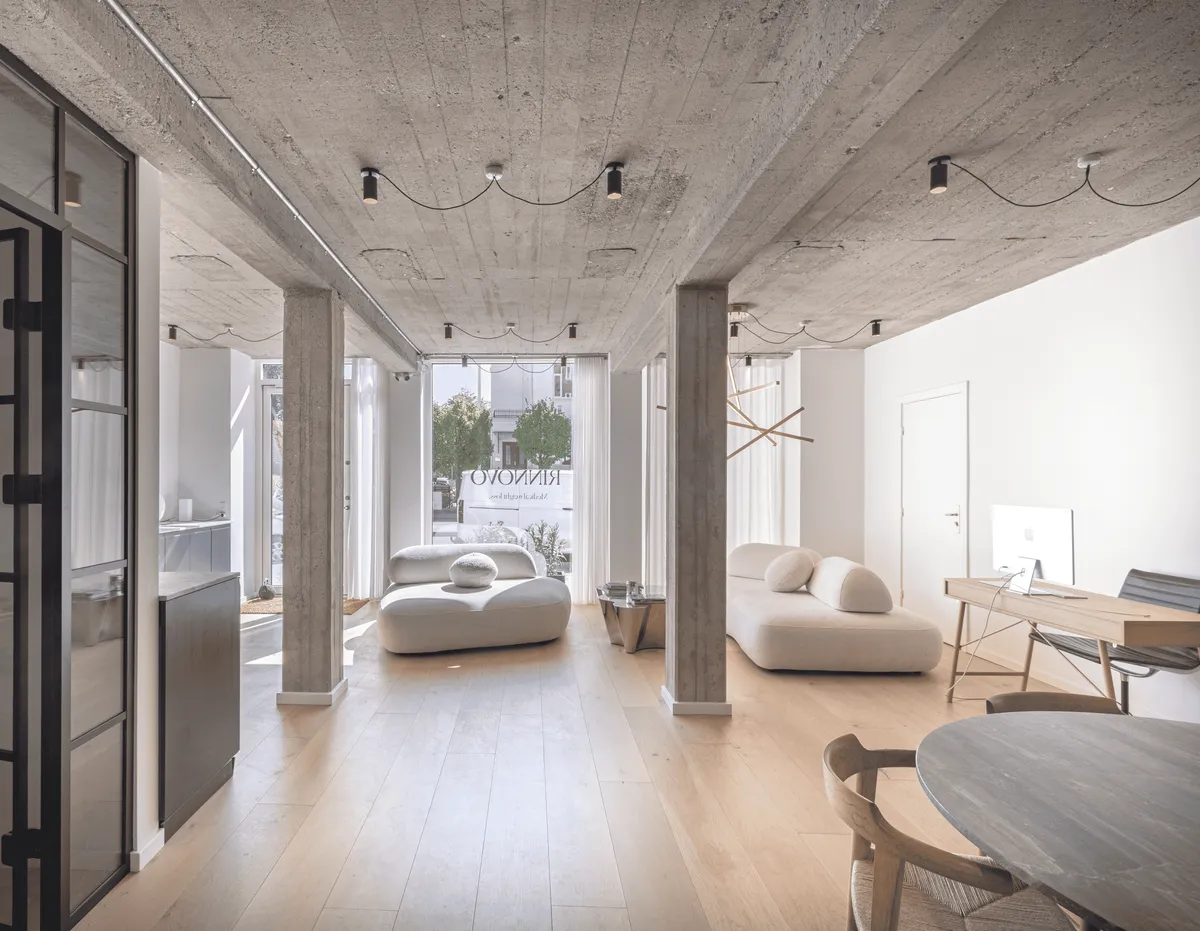 En stor og lys stue med højt loft, hvidt vægmaleri og lysegult gulv. Der er to komfortable sofaer og et skrivebord med monitor, stol og lyse lysekroner