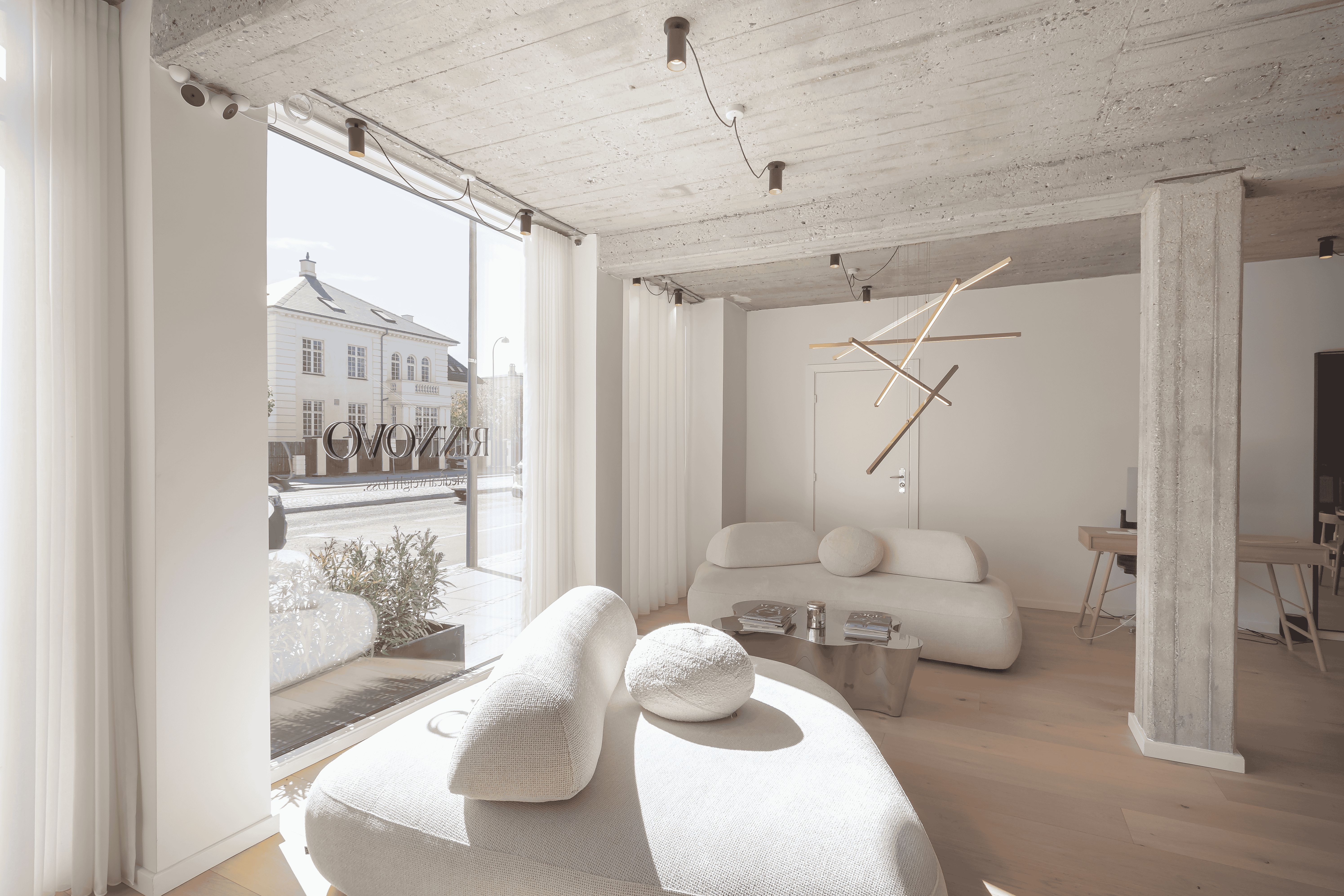 Moderne og minimalistisk interiør med hvide vægge, møbler og højloft. Lyse loftslampe og elegant sofa med kuglepuder. Glasdør og trægulv.