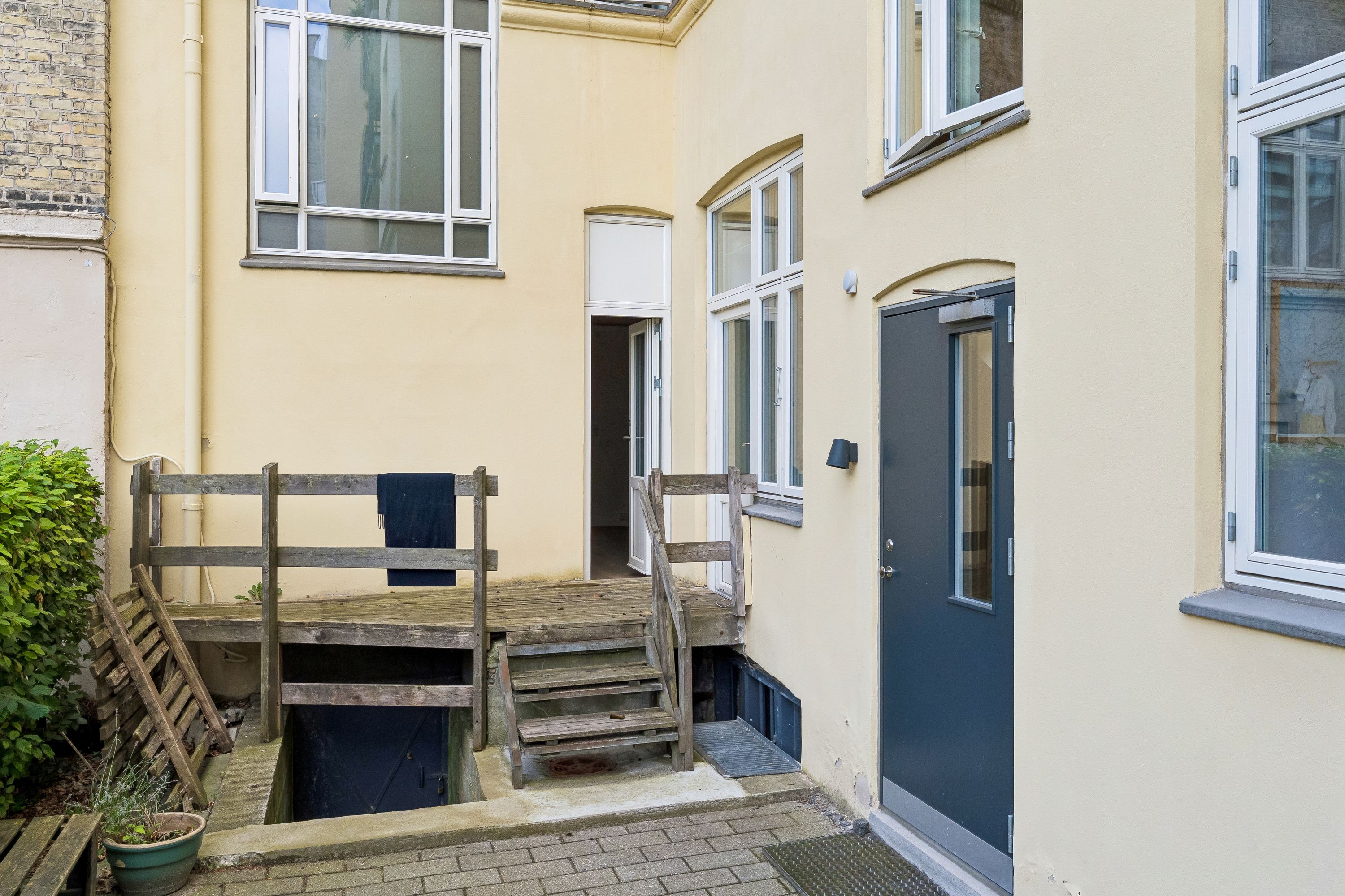 Gult byggede lejlighedshus med tre vinduer og en dør til højre, terrasse med trætrapper og stiger og en hængende tøjstativ med en blå trøje