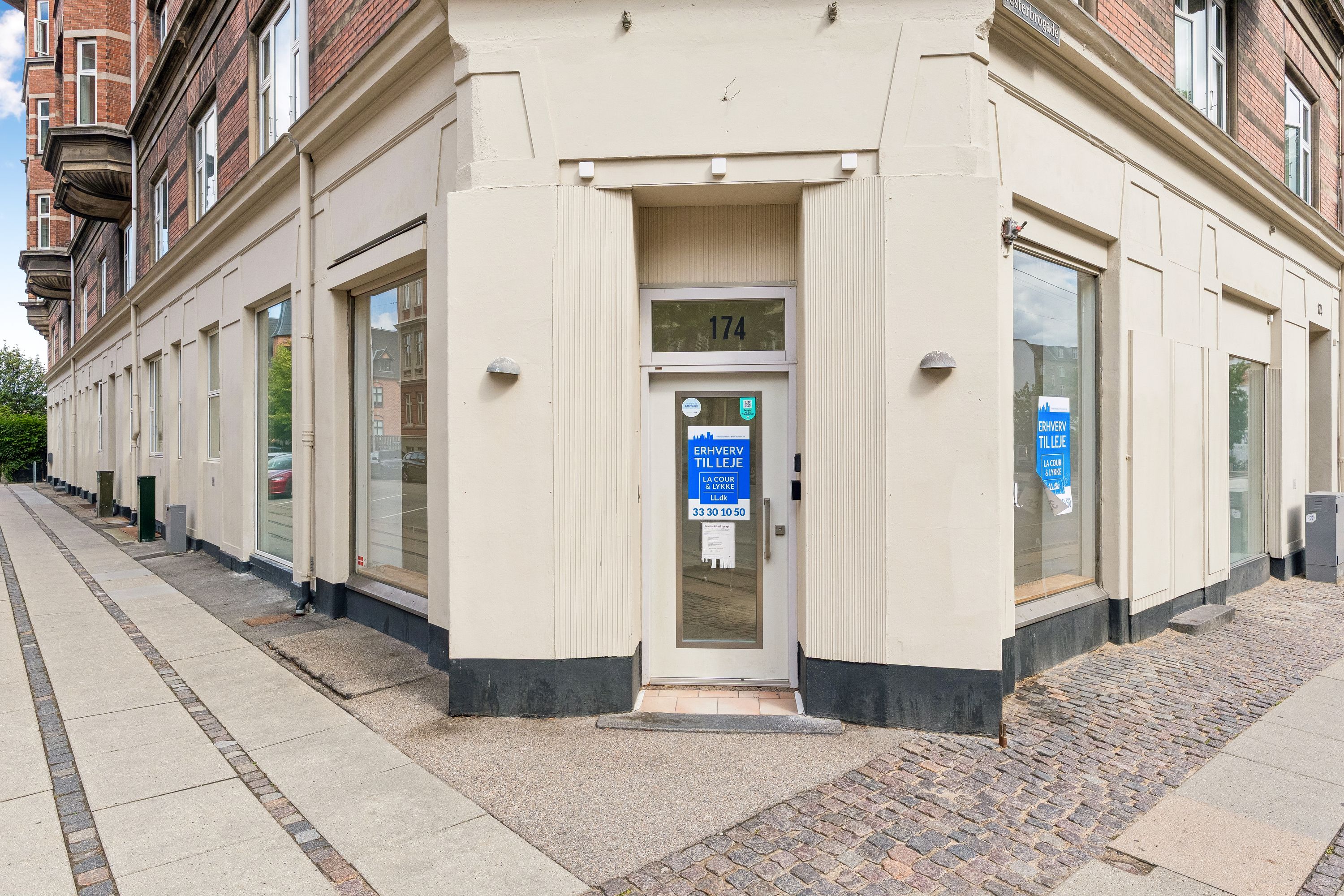 Foto af en butiksfront med glasvinduer, hvor der står "Ervvær til leje" på dørens glas. Der er også et skilt med "La Cour & Lynge" og en telefonnummer på døren.
