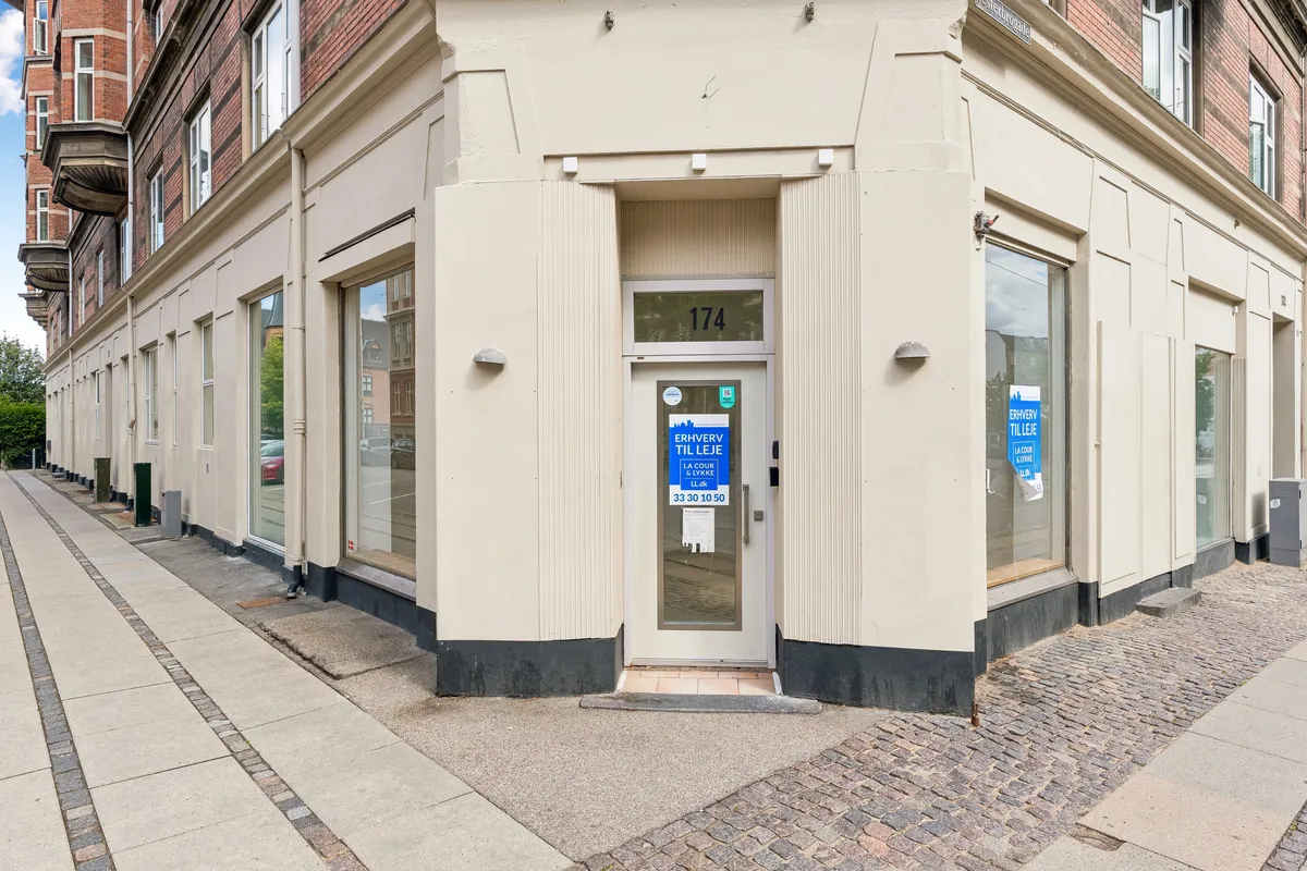 Foto af en butiksfront med glasvinduer, hvor der står "Ervvær til leje" på dørens glas. Der er også et skilt med "La Cour & Lynge" og en telefonnummer på døren.