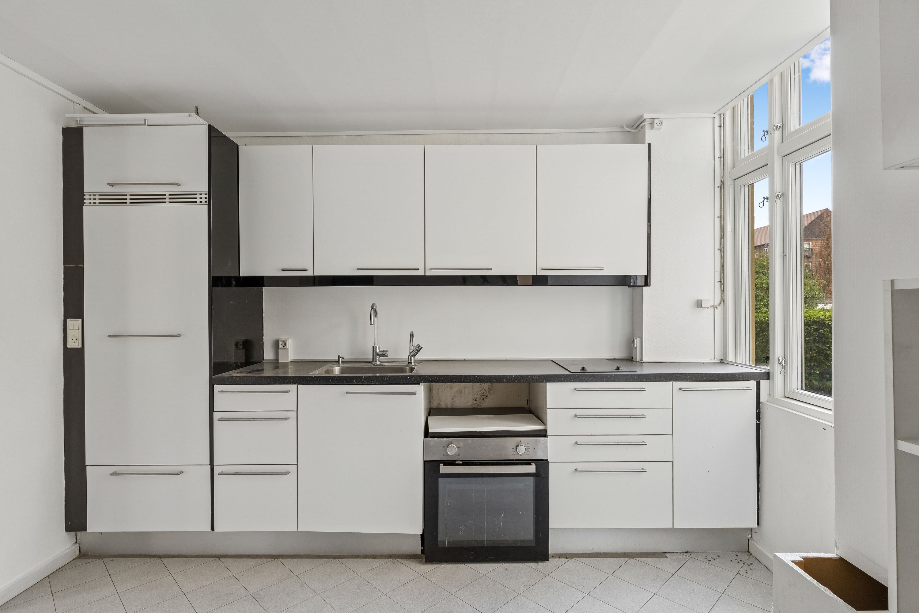 Rent køkken med hvide skabe og gult gulv, sort bordplade og lysegrå loft. Højt vinduesparti med udsigt til gård og hus.
