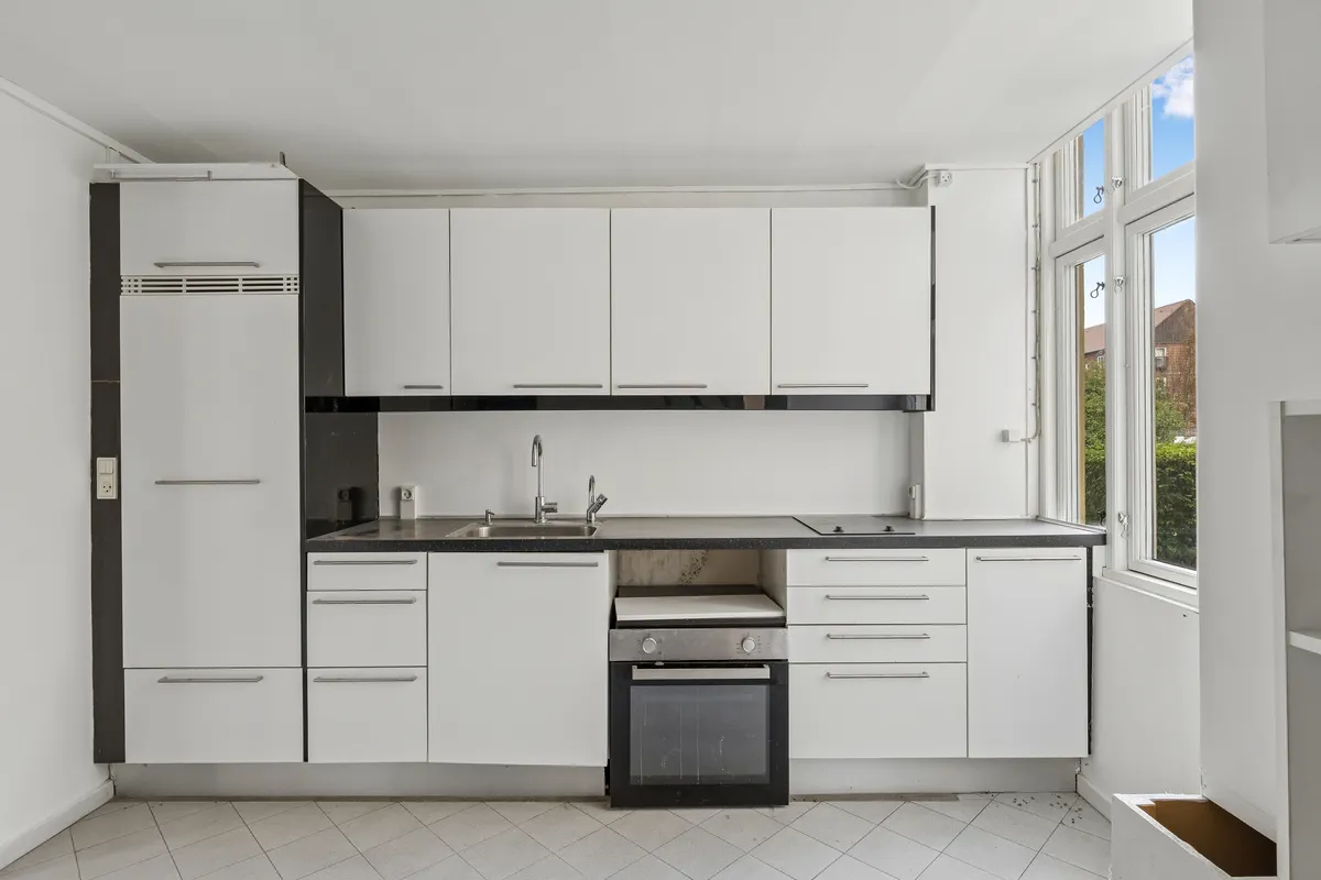 Rent køkken med hvide skabe og gult gulv, sort bordplade og lysegrå loft. Højt vinduesparti med udsigt til gård og hus.