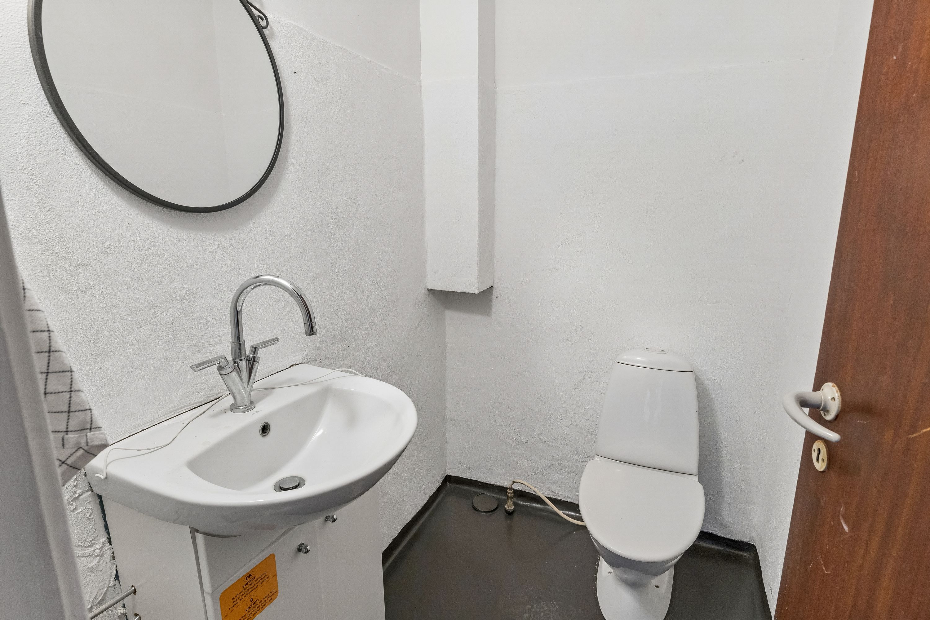 En hvid badeværelse med hvidt toilet, hvidt vasketøj, hvid væg og hvidt loft samt et rundt spejl med sort ramme