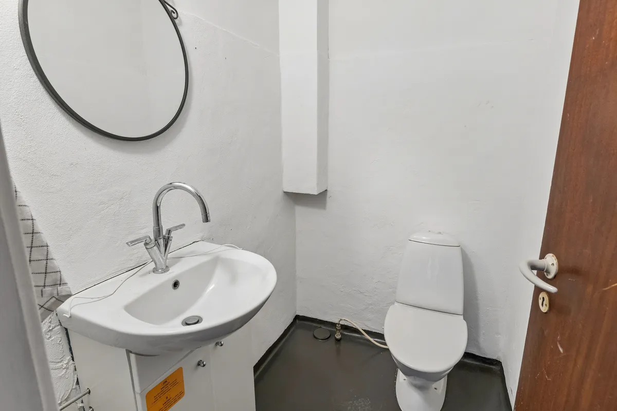 En hvid badeværelse med hvidt toilet, hvidt vasketøj, hvid væg og hvidt loft samt et rundt spejl med sort ramme