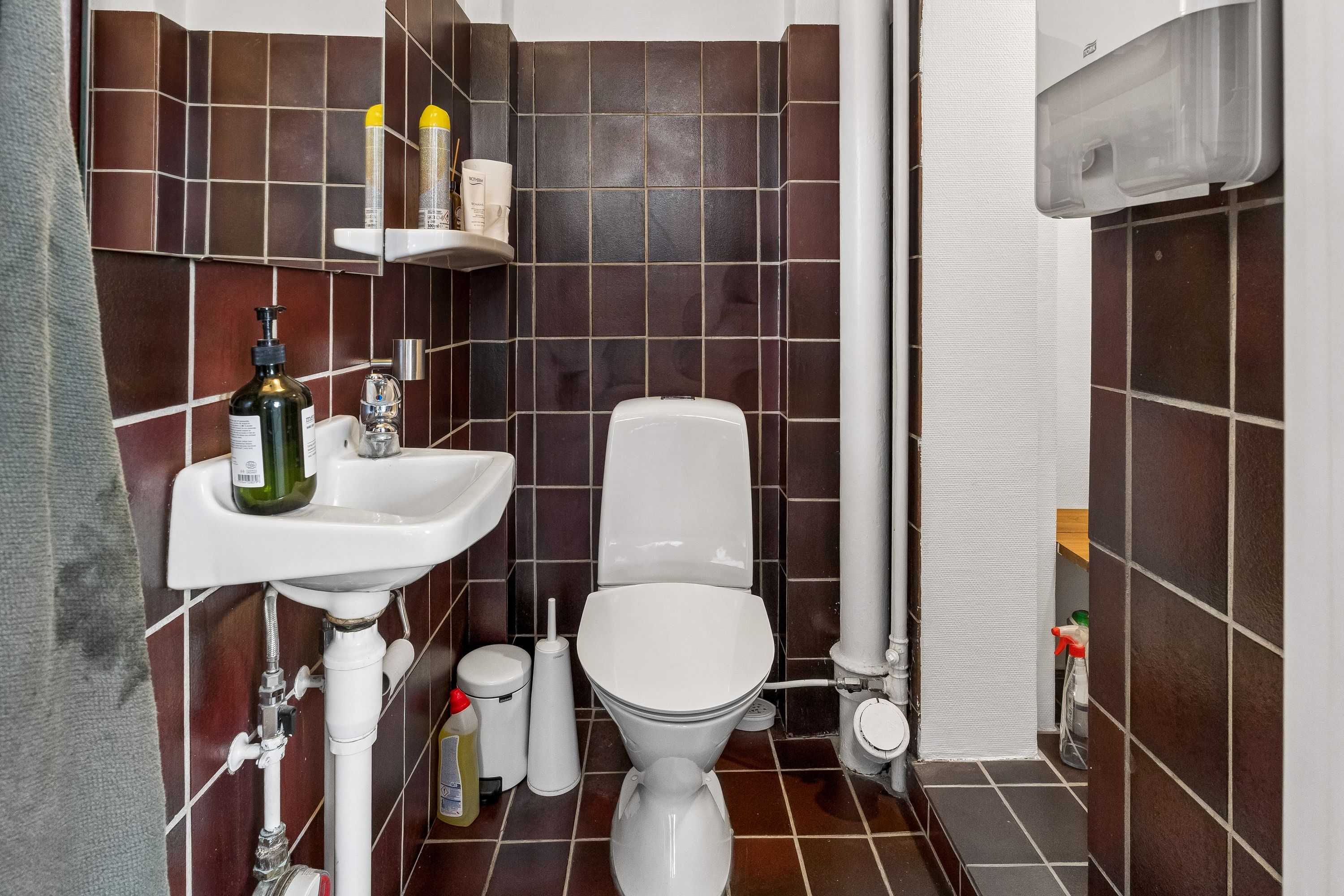 Lyst badeværelse med gulv- og vægfliser, hvidt toilet og håndvask med sæbe, toiletpapir og desinfektionsmiddelflaske samt toiletbørste