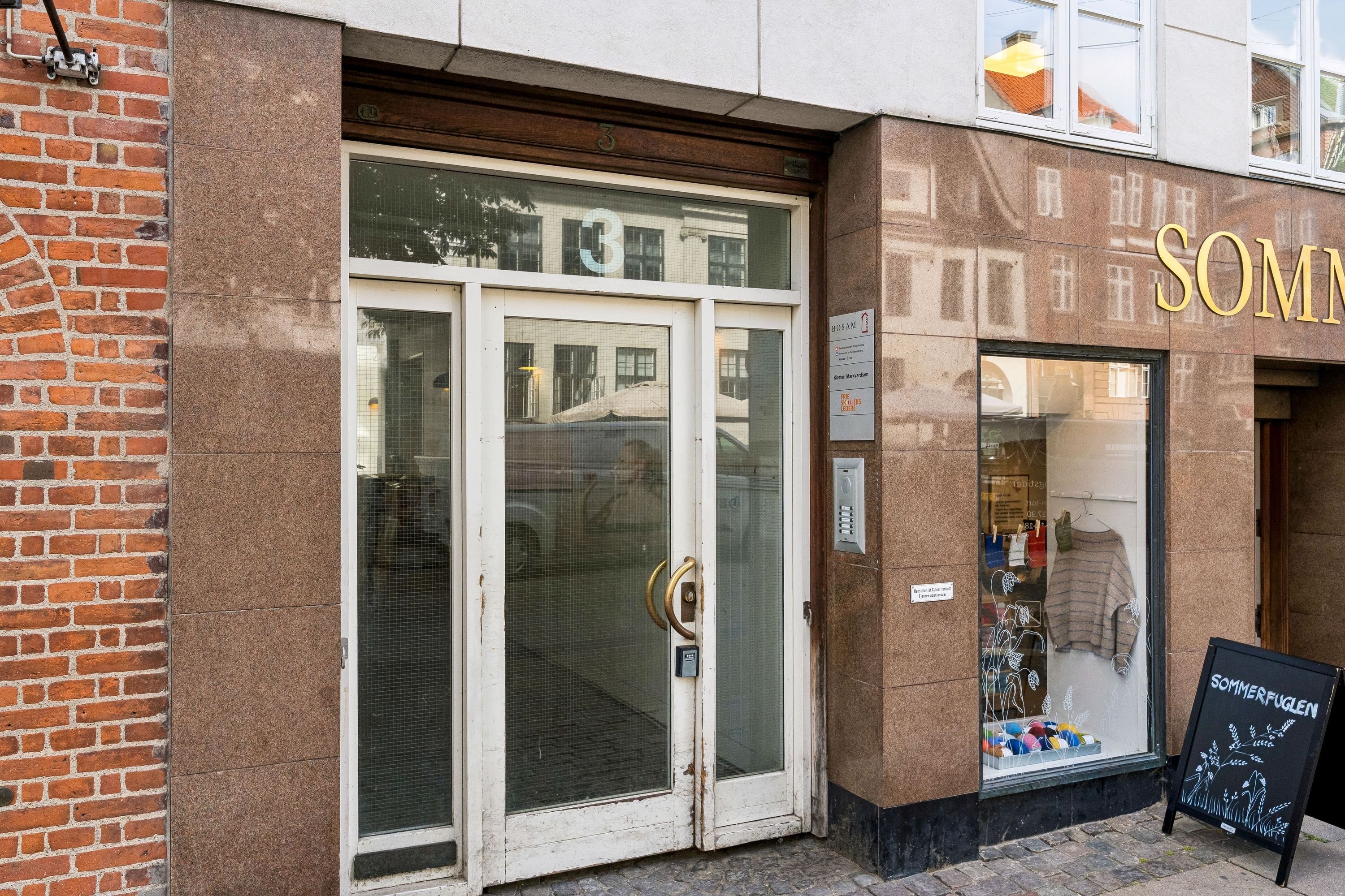 Kontor til leje på Vandkunsten 3, 1., 1467 København K med glasdøre og guldbeslag. En sort skiltstav placeret til højre for døren og et vindue med teksten Sommerfuglen