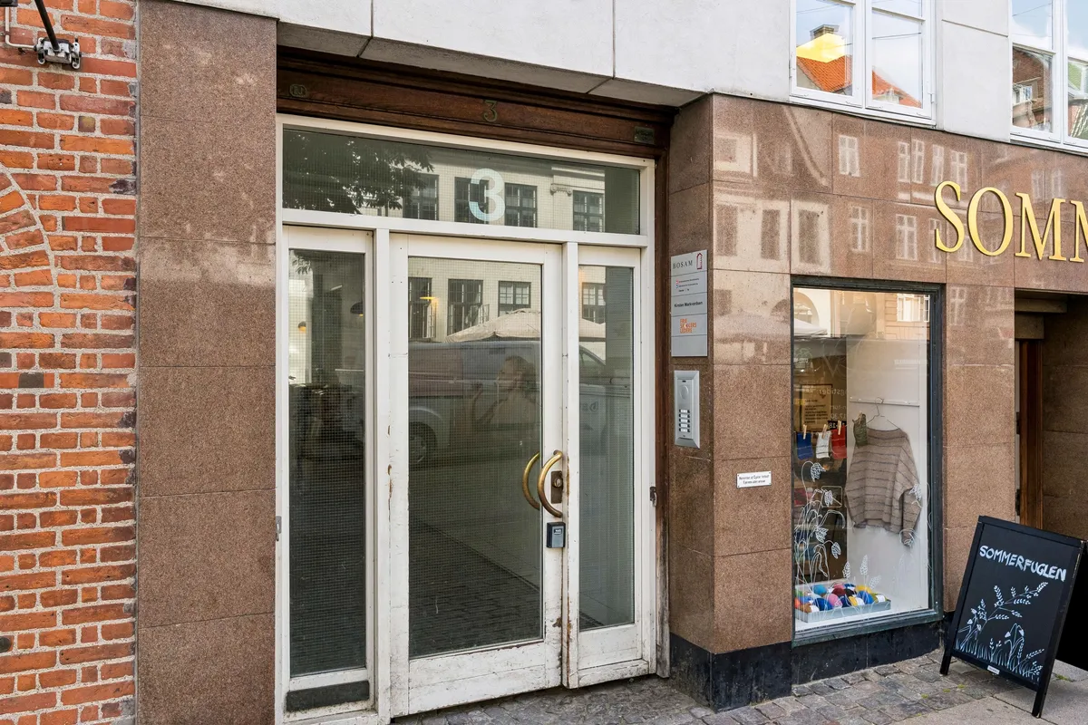 Kontor til leje på Vandkunsten 3, 1., 1467 København K med glasdøre og guldbeslag. En sort skiltstav placeret til højre for døren og et vindue med teksten Sommerfuglen