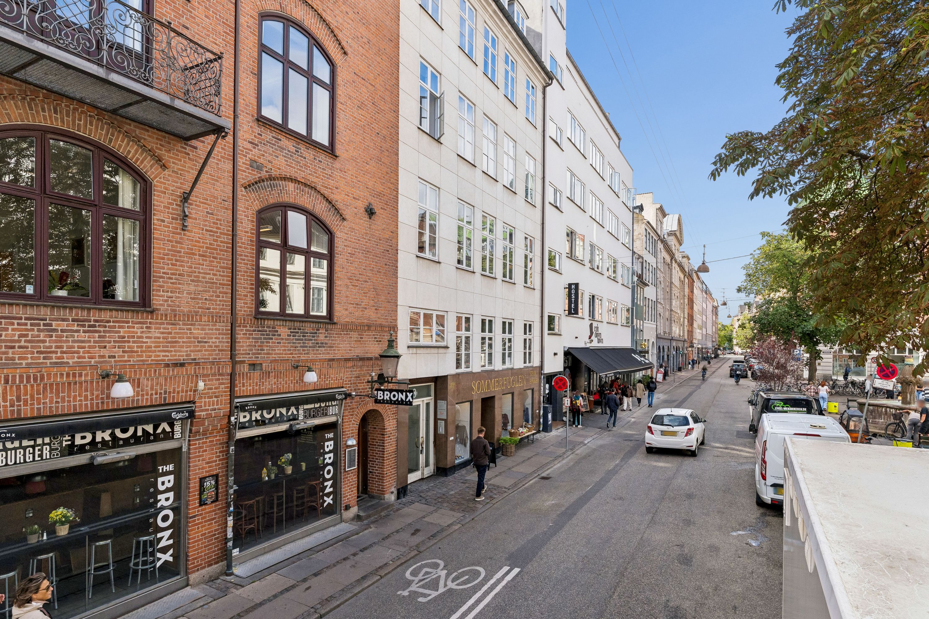 Kontor med glasvinduer på Vandkunsten 3, København K. Biler parkeret langs vej, folk vandrer, grønne træer og et stop-skilt
