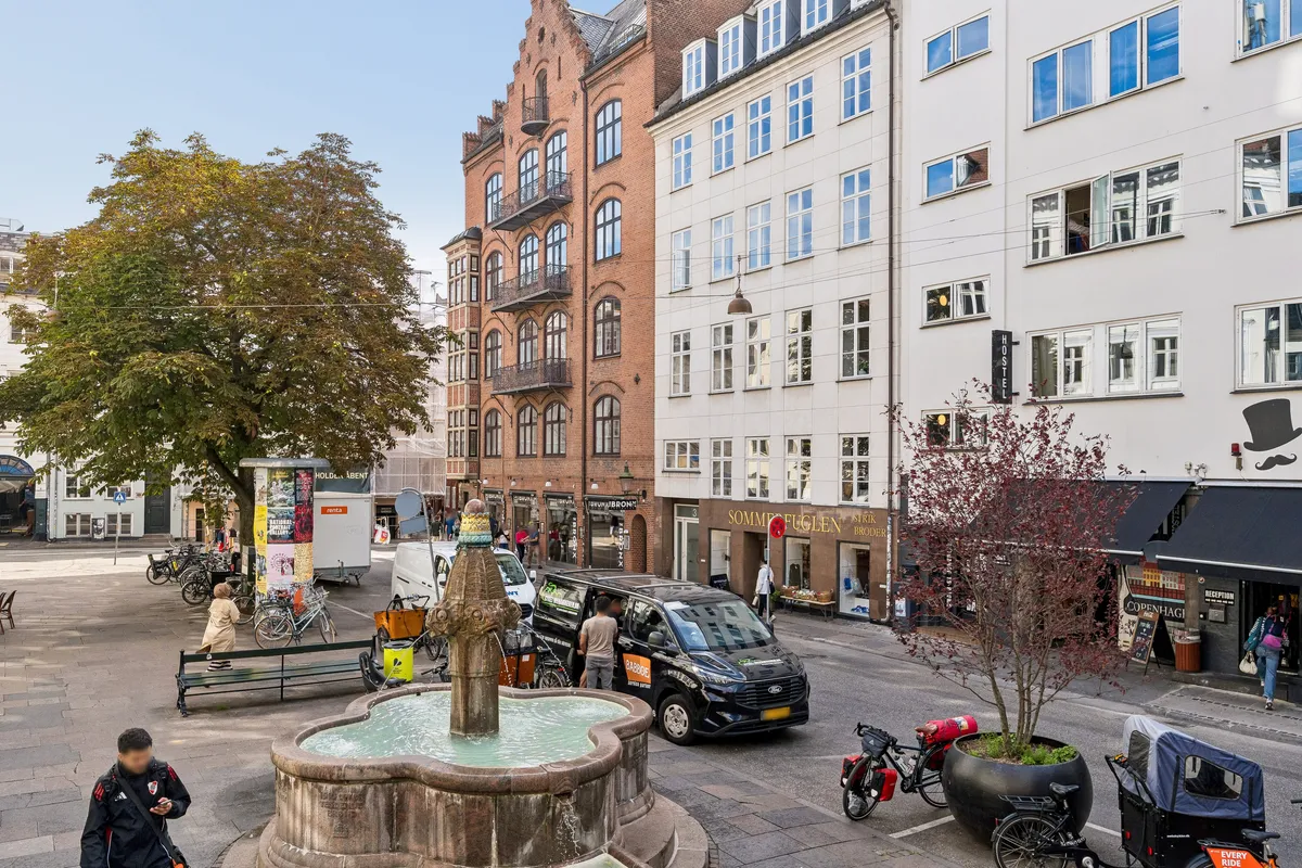 Et kontor på Vandkunsten 3 i København K med parkeringsmuligheder og et vandfadsgrav. Folk går og cykler forbi, og der er flere biler parkeret. En kvinde går og snakker med en mand ved en bil.