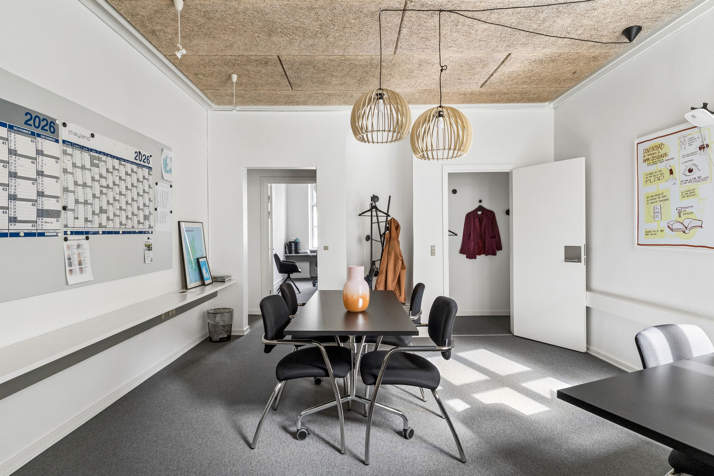 Lys kontorrum med højt loft og lyse gulve. Rundt bord med stole, et loftsbord og loftslampe. Hvid væg med planlægningskalender og noter.
