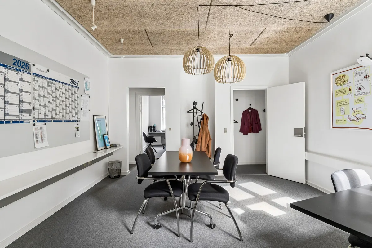 Lys kontorrum med højt loft og lyse gulve. Rundt bord med stole, et loftsbord og loftslampe. Hvid væg med planlægningskalender og noter.