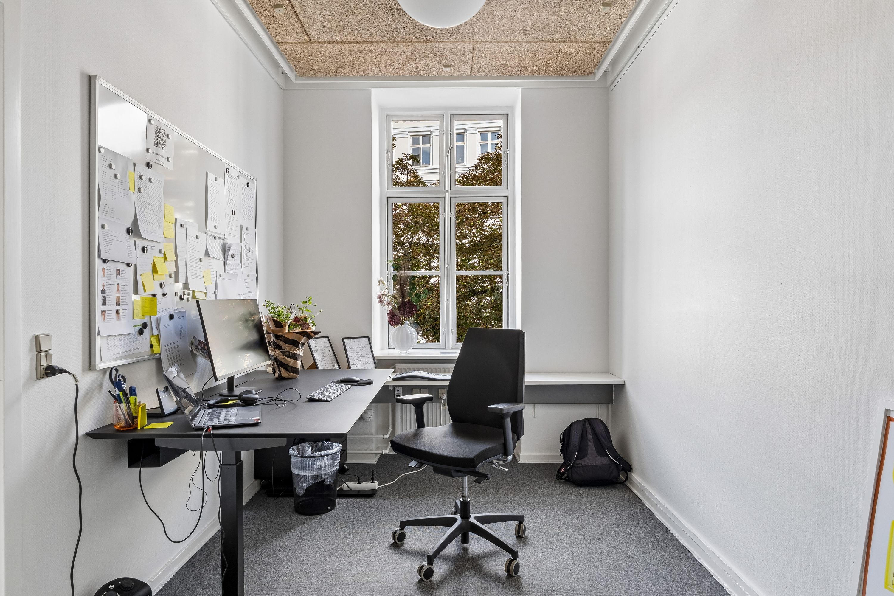 Kontor med minimalistisk design og hvidt interiør. Bord med computer, tastatur og mus. Sort stol med hjul og bagage på gulvet. Magnetbræt med papirer og noter. Vindue med udsigt til grønne træer.