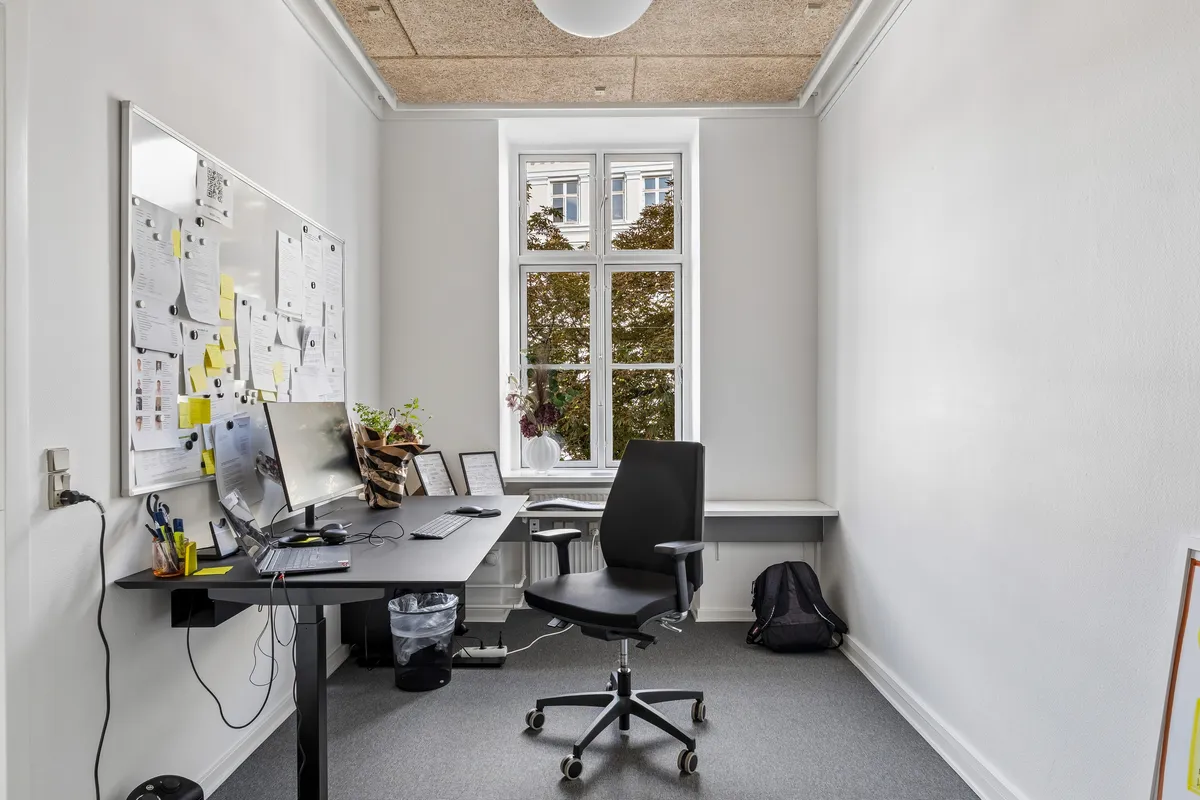 Kontor med minimalistisk design og hvidt interiør. Bord med computer, tastatur og mus. Sort stol med hjul og bagage på gulvet. Magnetbræt med papirer og noter. Vindue med udsigt til grønne træer.