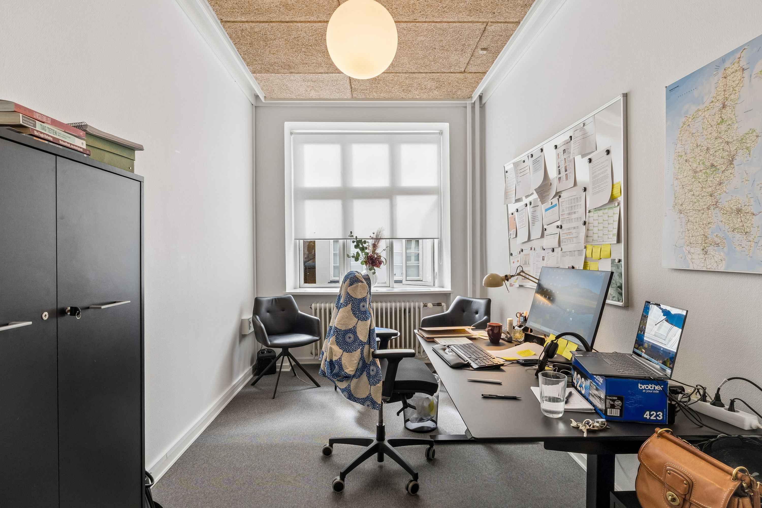 Kontor med skrivebord, computere og stol. Der er et bibliotek i hjørnet og et landkort på væggen.
