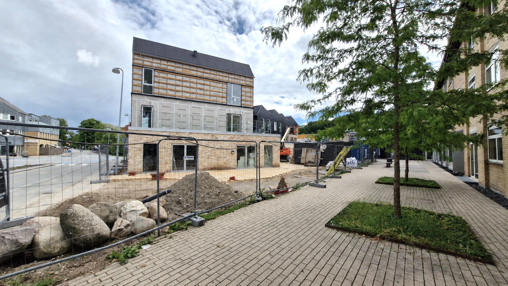 Under konstruktion er en ny bygning med terrasser og vinduer i Allerød på Frederiksborgvej. Der er en høj mur og sten ved vejkant og konstruktionsmaterialer.