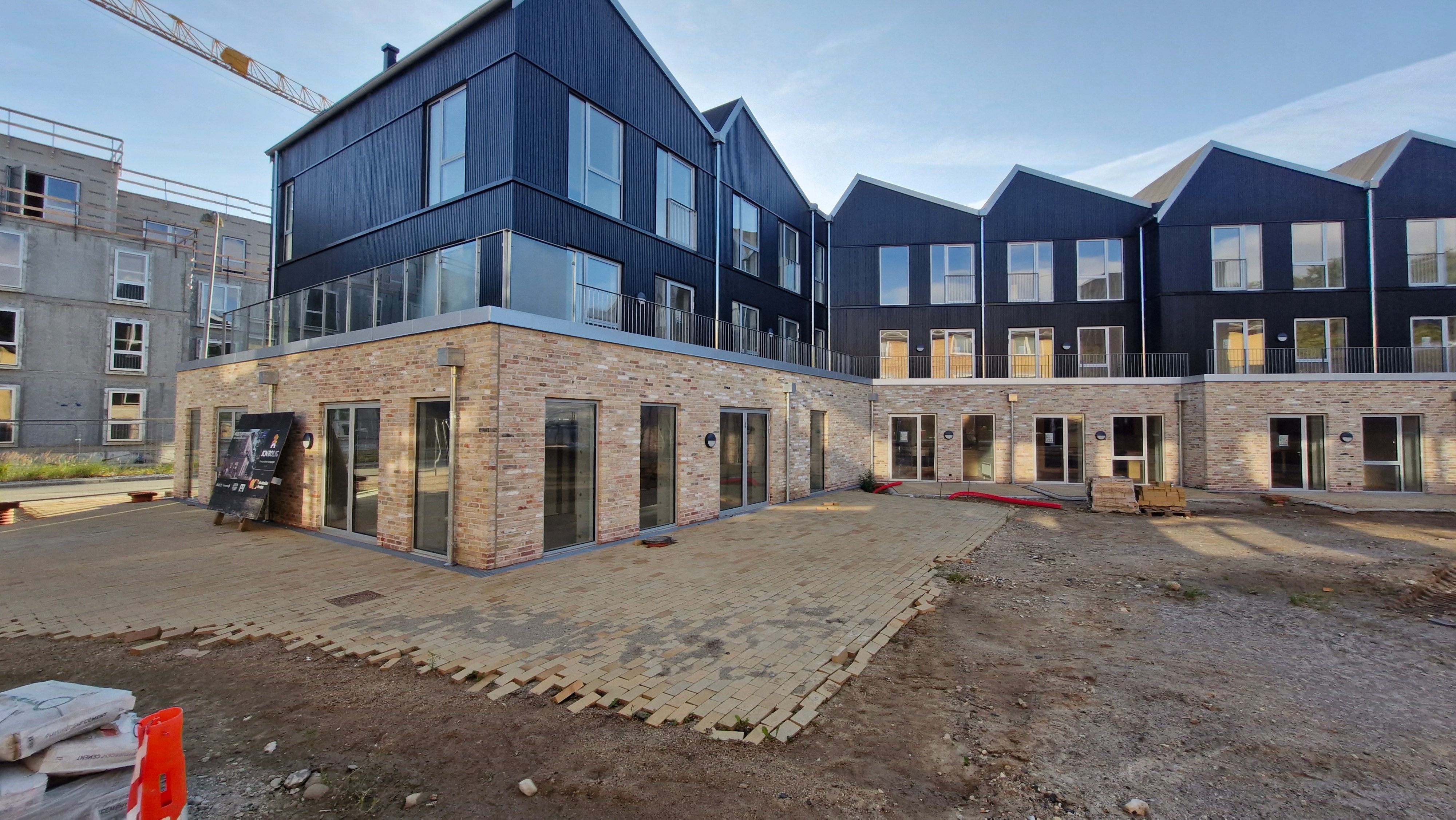 Moderne hus med skrånende tag og glasvinduer. Bygningen er i fase af opførelse med røde mursten og glas. Bagved bygningen er en skur og et højt kran.