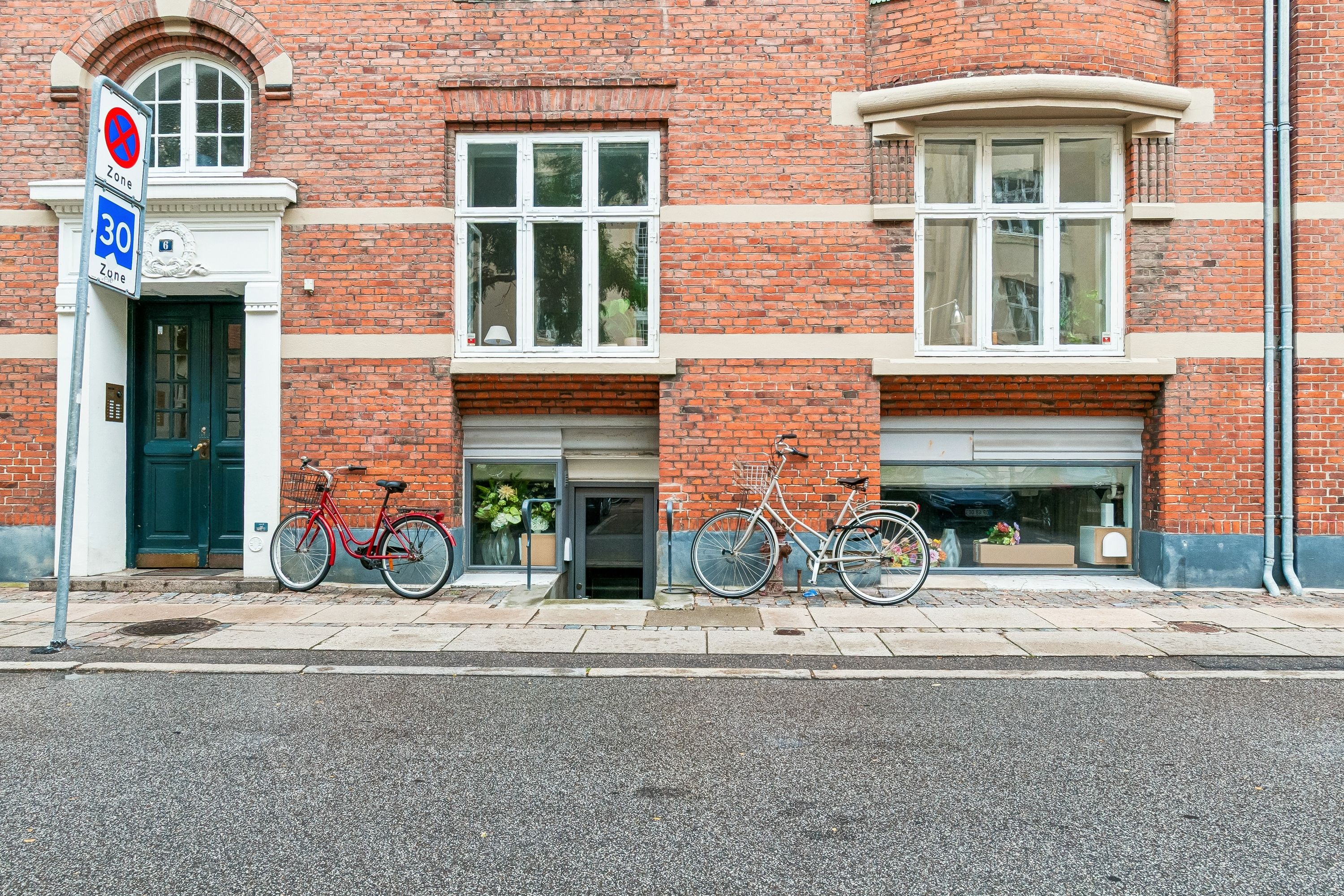 To røde og en hvid cykel parkeret foran et rødt murstenshus med store glasruder og et blåt 30-zoneskilt på gaden