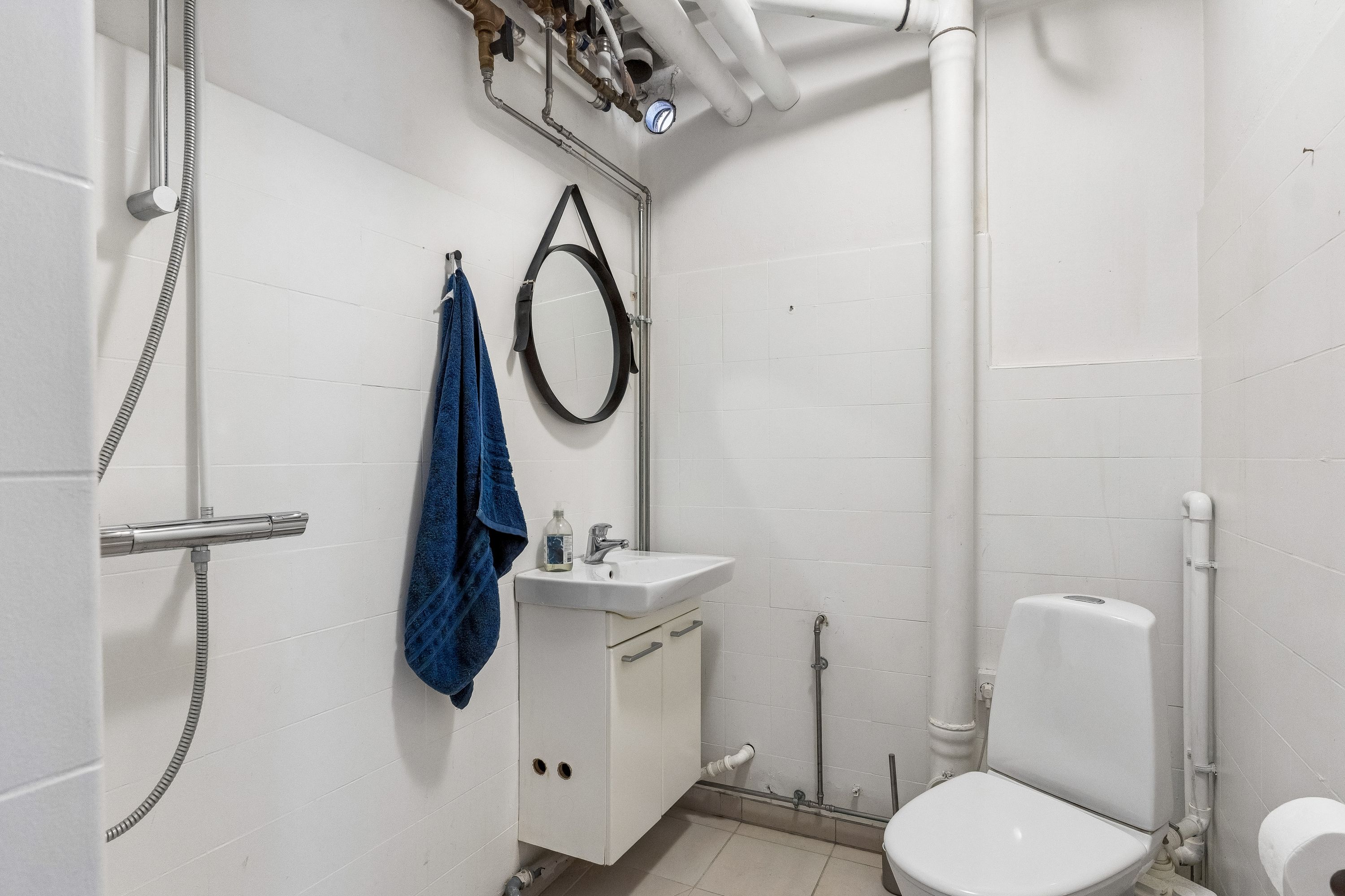Foto af et lille badeværelse med en hvid badeværelseskabinet, toilet, håndklæde, spejl og en vandhane med brusebad. Det er et minimalistisk og velholdt badeværelse med lyse, hvide vægge og praktiske opbevaringsmuligheder
