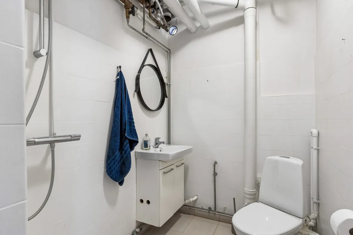 Foto af et lille badeværelse med en hvid badeværelseskabinet, toilet, håndklæde, spejl og en vandhane med brusebad. Det er et minimalistisk og velholdt badeværelse med lyse, hvide vægge og praktiske opbevaringsmuligheder