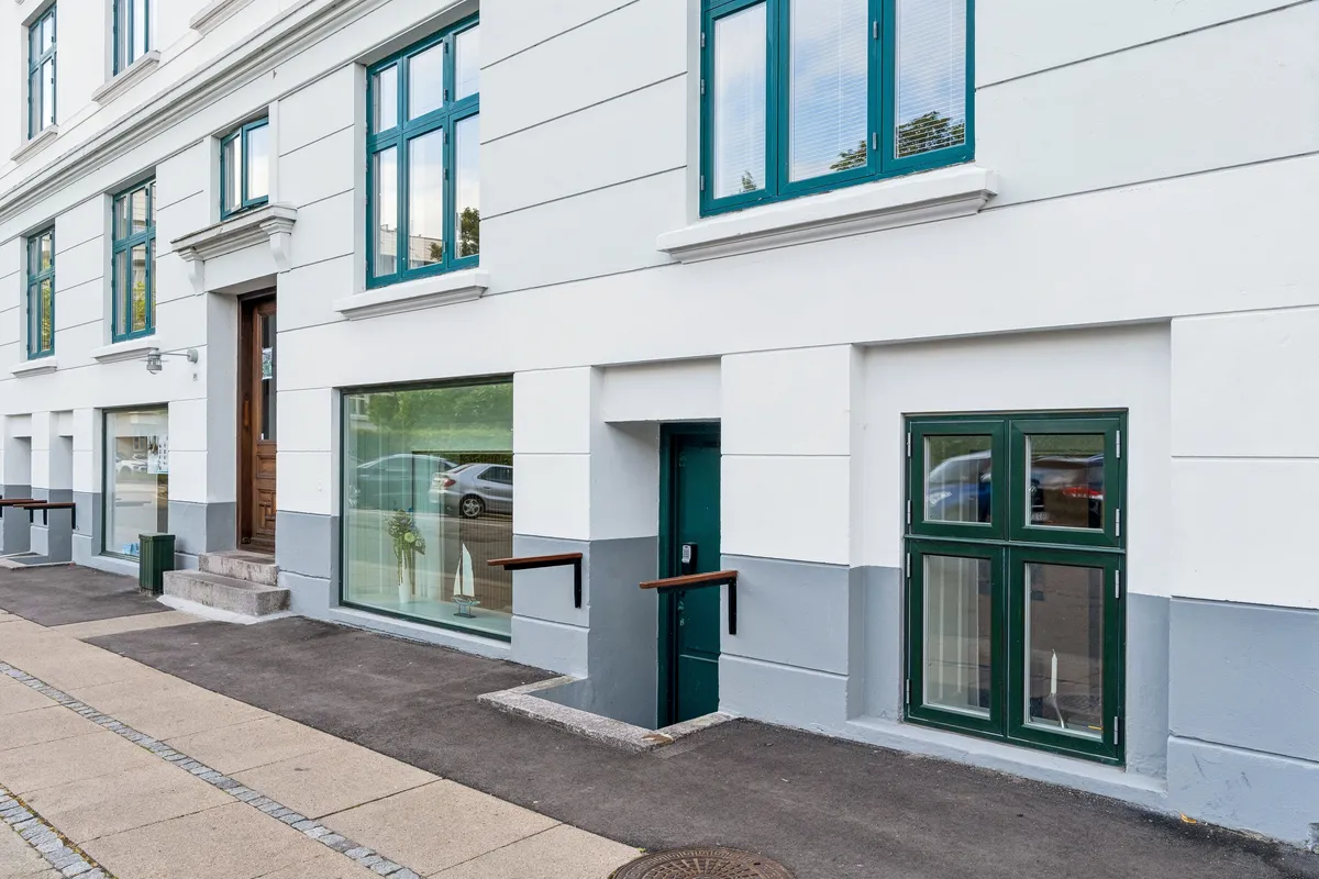 Kontor til leje på Næstvedgade 28 med lyse vinduerne og lysegrå væg. Der er to indgange og et gulvudstødningslukke. Der er skrånende vejforhold på gaden med asfalt.