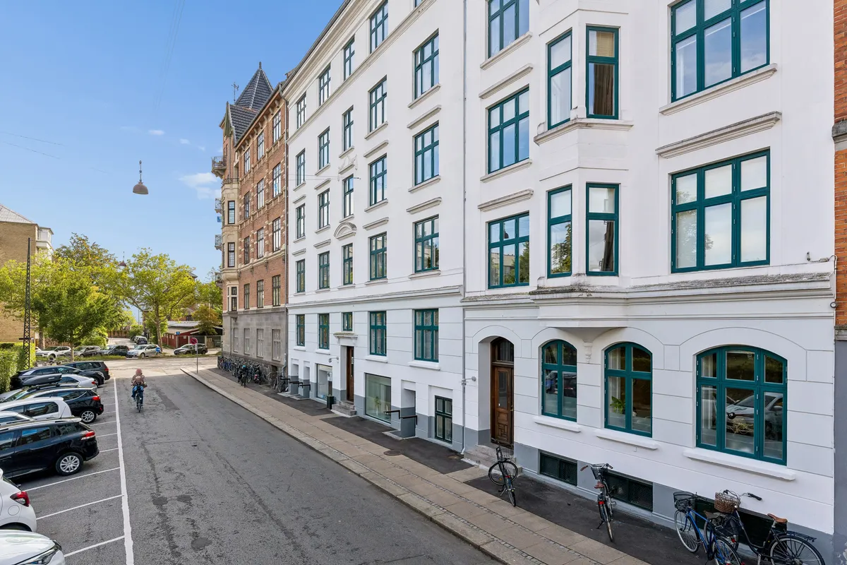 Kontorbyggeri med flere parkerede biler og cykler i forgrunden, hvor en cykelkørende passerer. Træet og facaden af bygningerne viser en rolig og fredelig gade.
