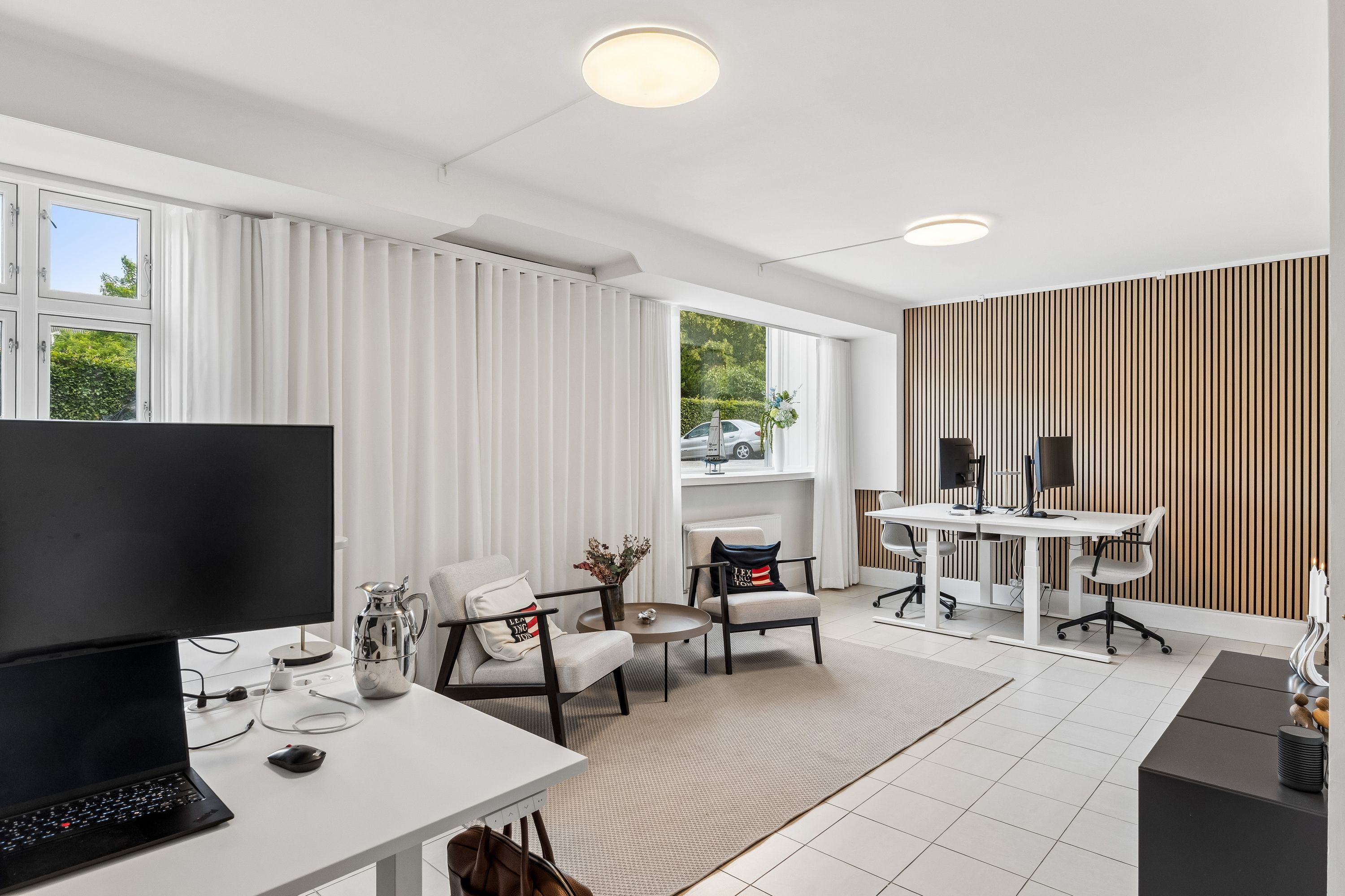 Kontorrum med moderne møbler og hvidt loft. To hvide skriveborde med computere og stolpe og en hvid sofa med pude og vase med blomster.