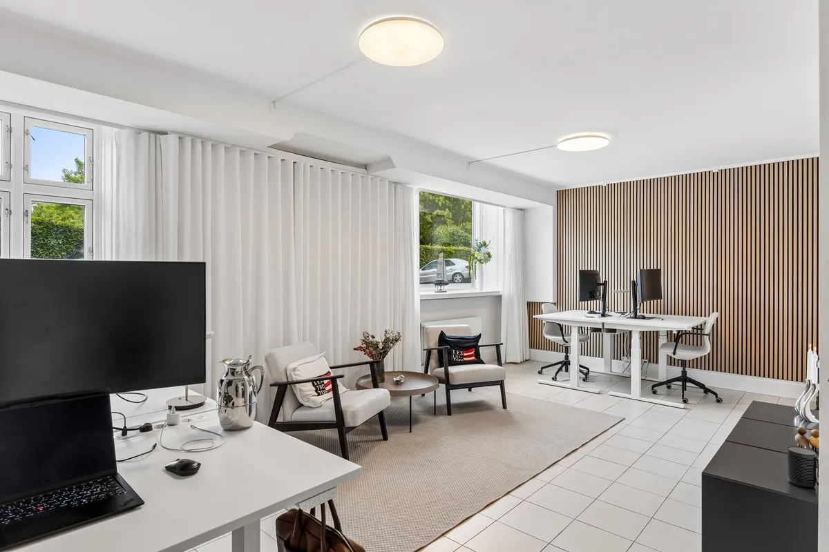 Kontorrum med moderne møbler og hvidt loft. To hvide skriveborde med computere og stolpe og en hvid sofa med pude og vase med blomster.