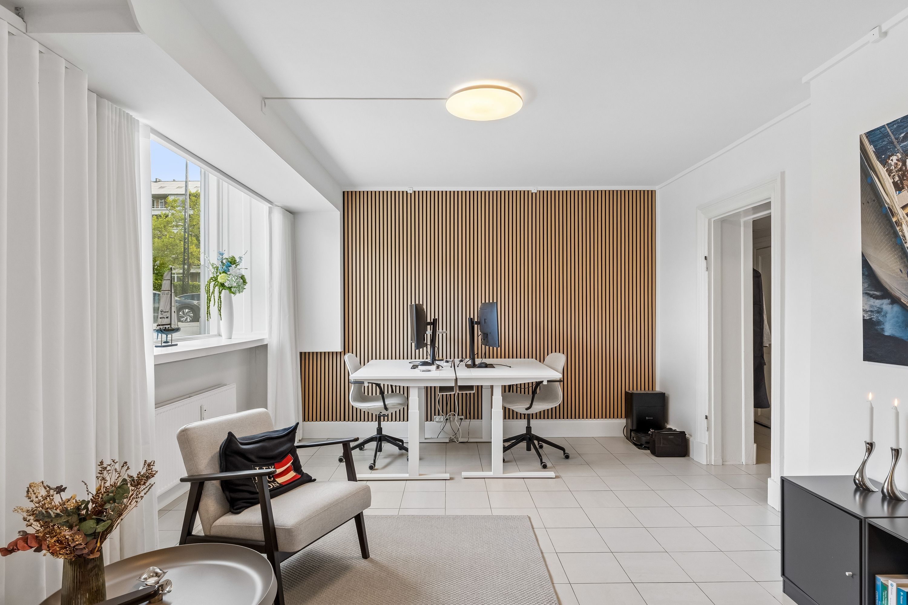Kontor med skrivebord og stole, lysekronen hænger fra loftet og træpanelet er på væggen. To monitorer og mus er på skrivebordet, der er en vase med blomster på vindueskarmen