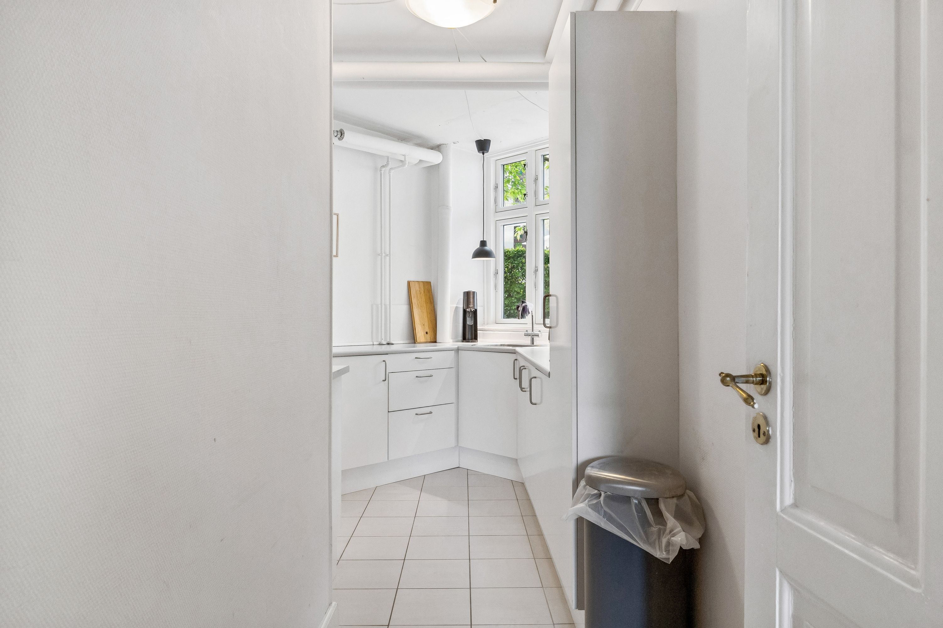 Et lyse og rummeligt køkken med hvide vægge og hvidt loft, høj kvalitet og moderne apparater samt en udsigt til gården med grønne planter og lyse vinduer