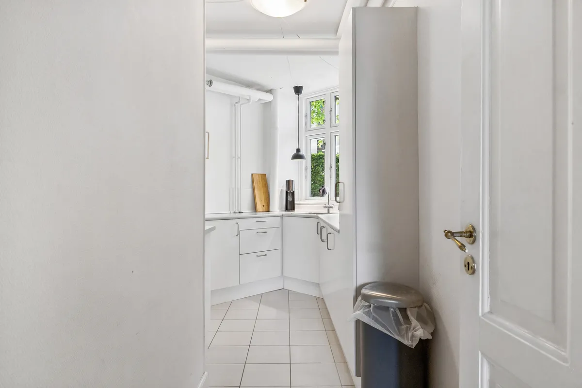 Et lyse og rummeligt køkken med hvide vægge og hvidt loft, høj kvalitet og moderne apparater samt en udsigt til gården med grønne planter og lyse vinduer